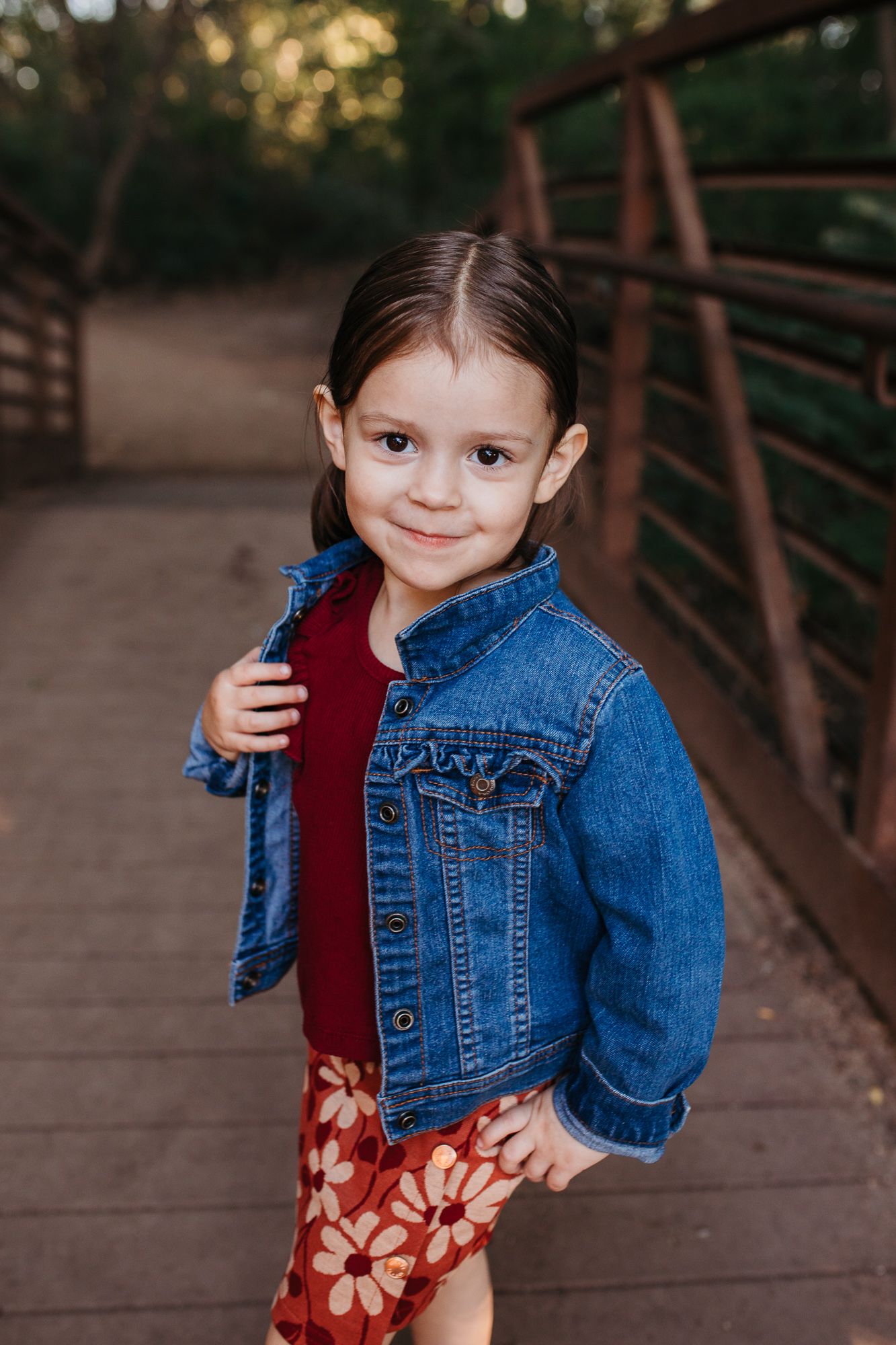 A child in a denim jacket and floral dress poses on a wooden bridge during golden hour -annual milestone photos Eagle Idaho