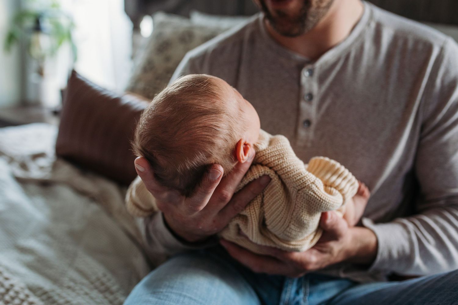 A parent holds a sleeping newborn baby wrapped in a beige sweater while sitting on a bed at home - lifestyle in-home newborn photos Boise