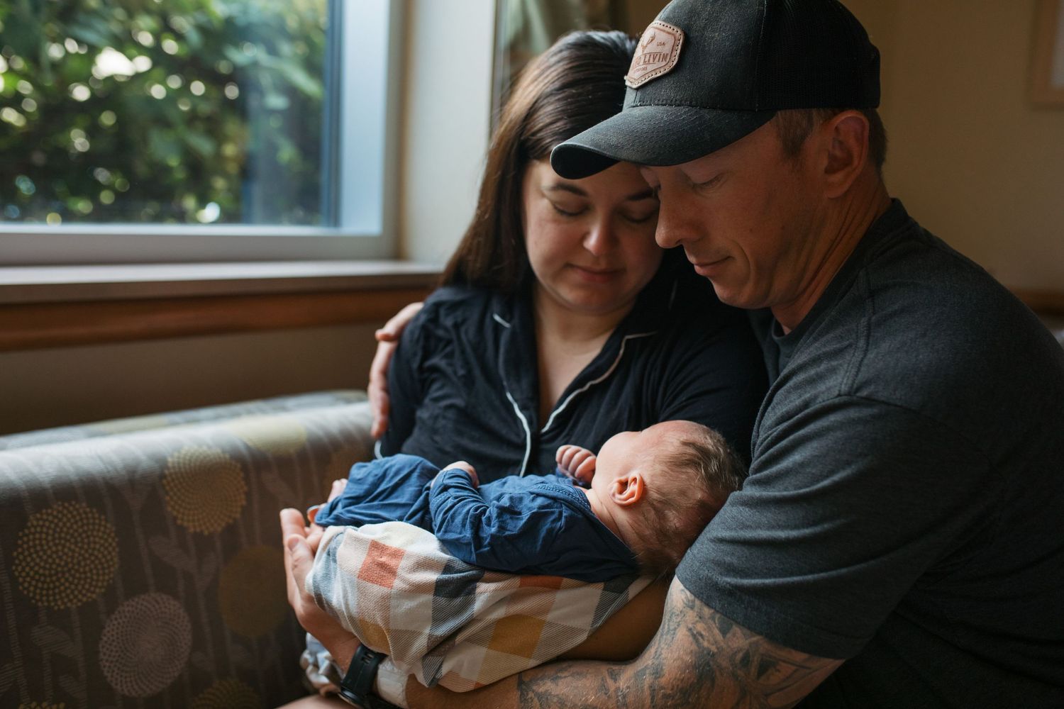 Two adults sit closely together on a couch while cradling a sleeping newborn baby wrapped in cozy blankets. Fresh 48 session Boise