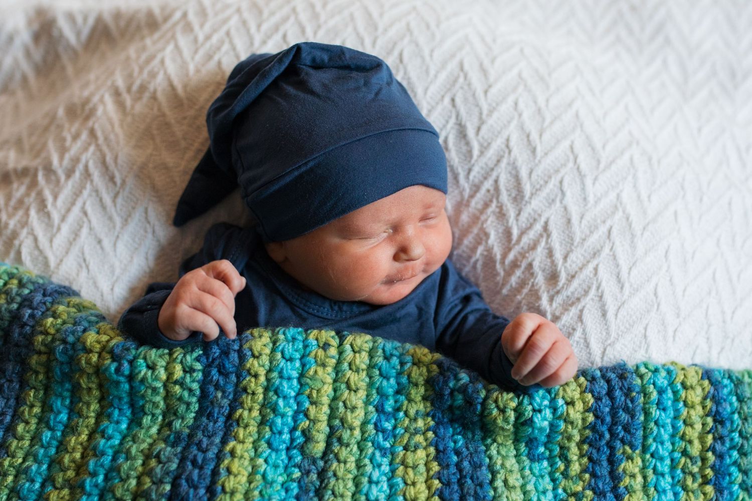 A baby wearing a blue hat sleeps peacefully under a colorful striped crocheted blanket - Fresh 48 session in hospital Boise, Idaho