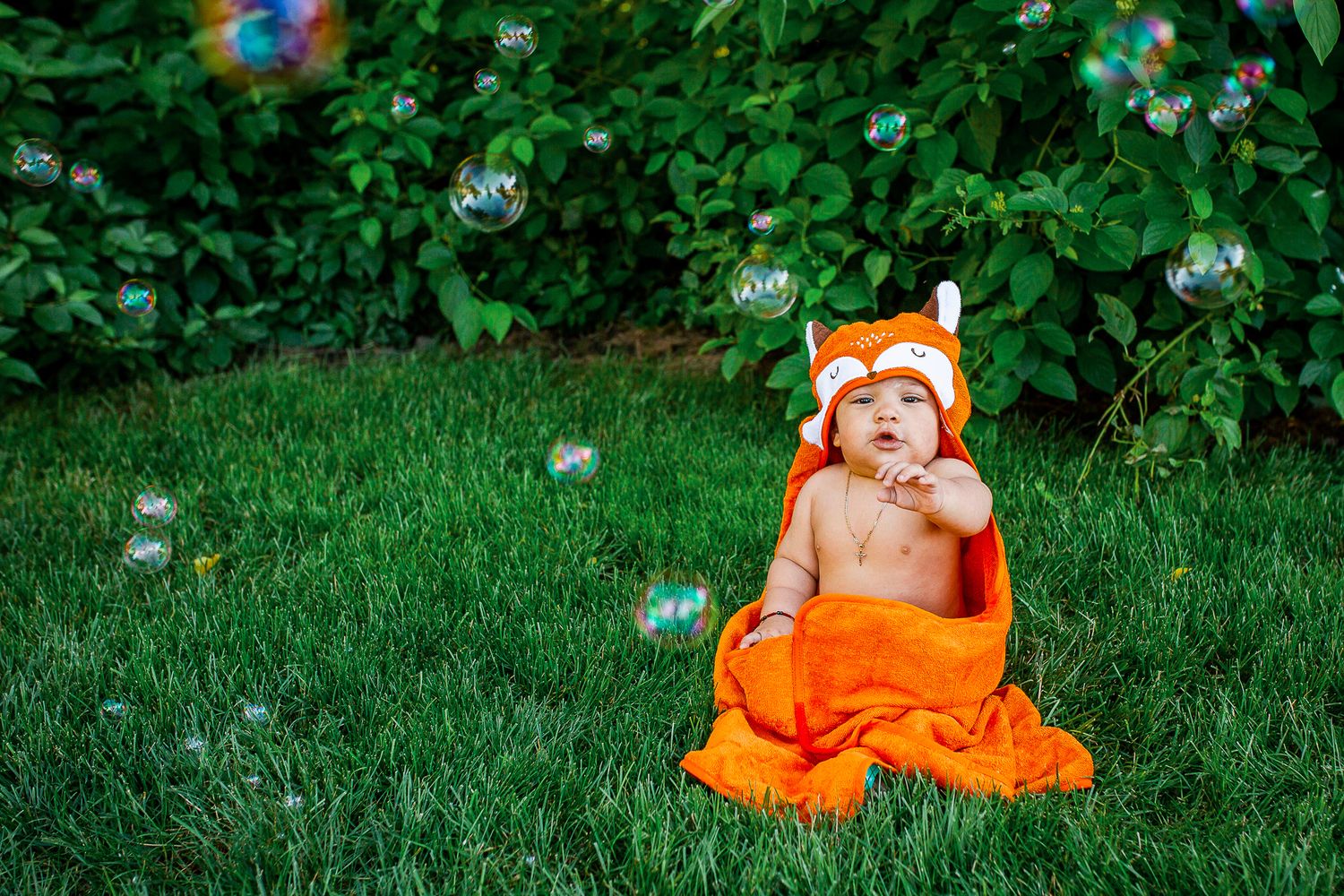 A baby wearing an orange fox costume sits on green grass while playing with soap bubbles floating in the air - 9 month milestone photos