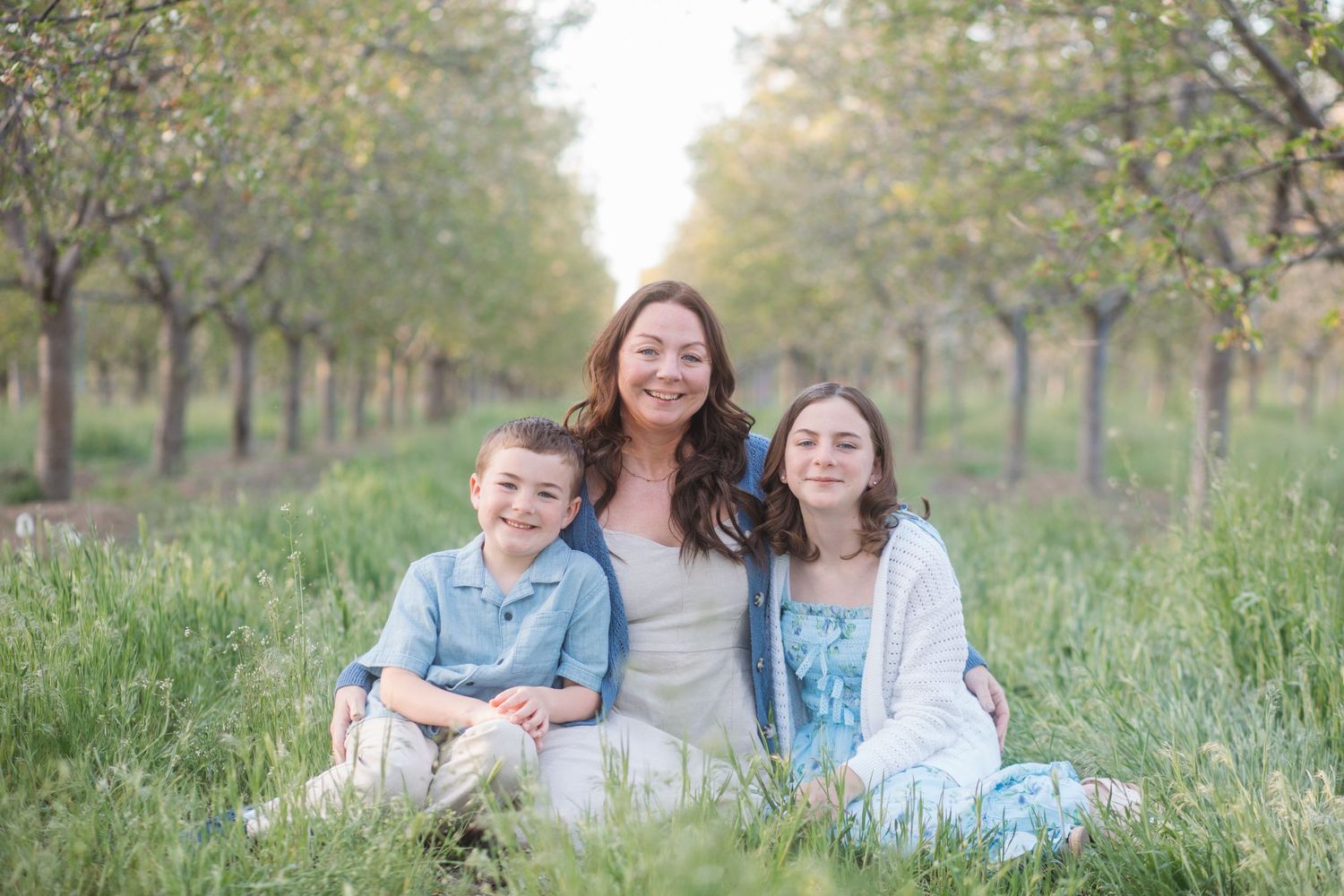 Mother and two kids sitting in grass under cherry blossom orchard during spring family session in Caldwell Idaho