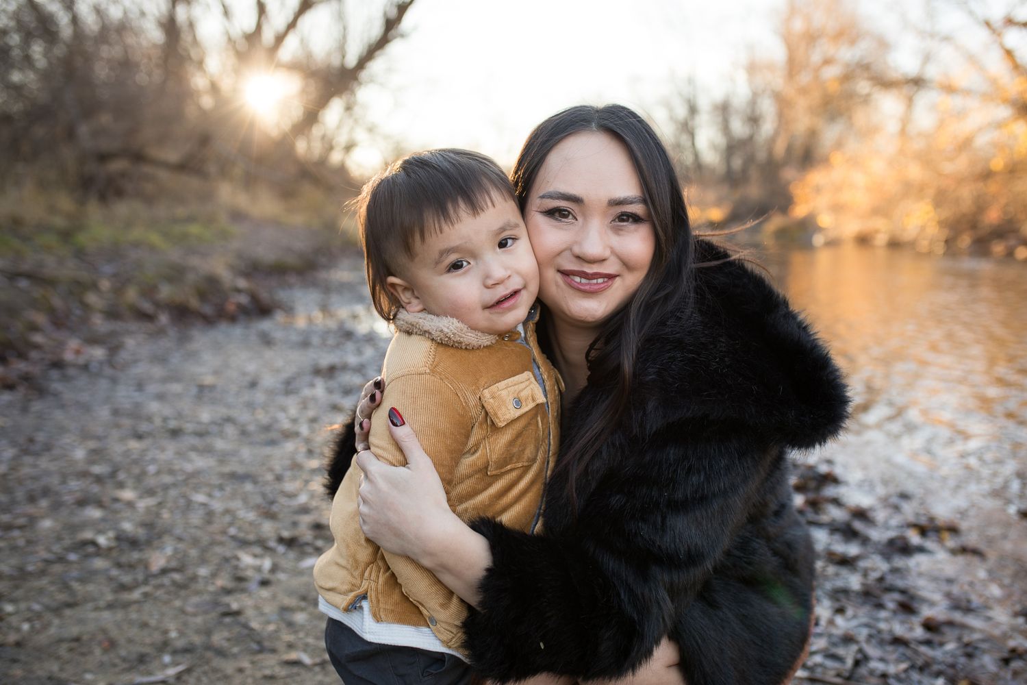 Mother hugging her toddler beside the river at Reid Merrill Park in Eagle, Idaho during golden winter sunset.