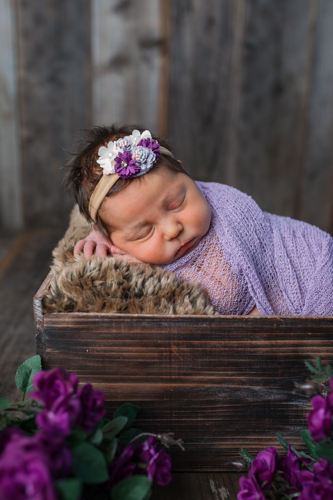 A sleeping infant wrapped in purple knit swaddle rests on wooden crate surrounded by vibrant purple flowers  studio newborn photography