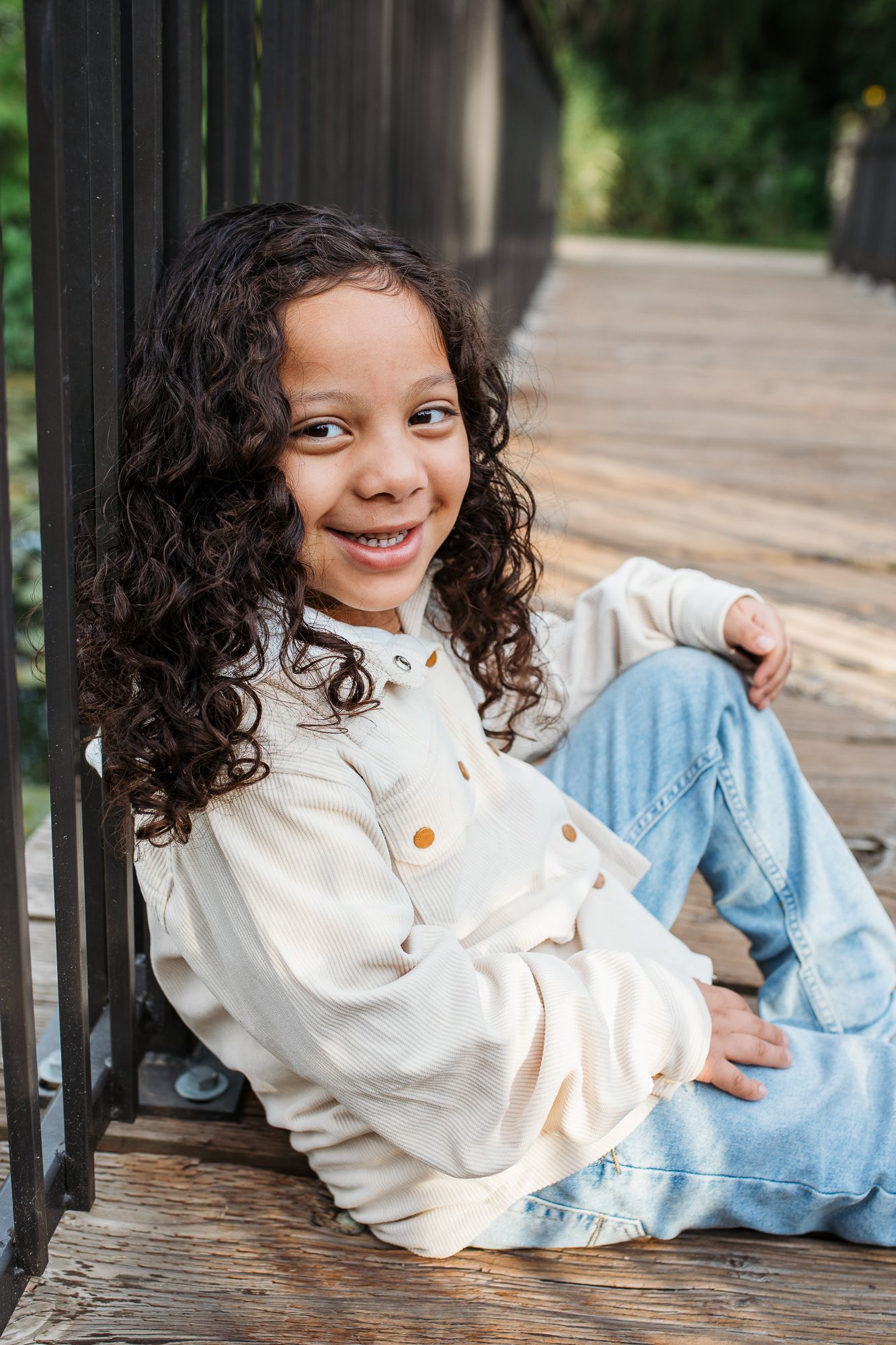 A child in a white sweater and jeans relaxes on wooden decking while showing a bright smile - birthday milestone photos Boise Idaho