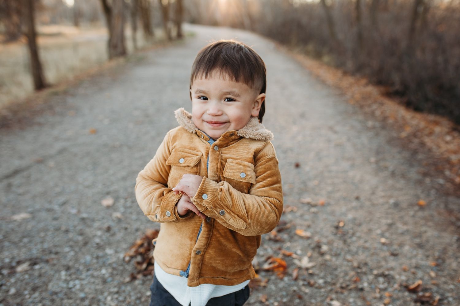A child wearing a brown coat plays with fallen autumn leaves on a forest path - lifestyle family photos in Boise
