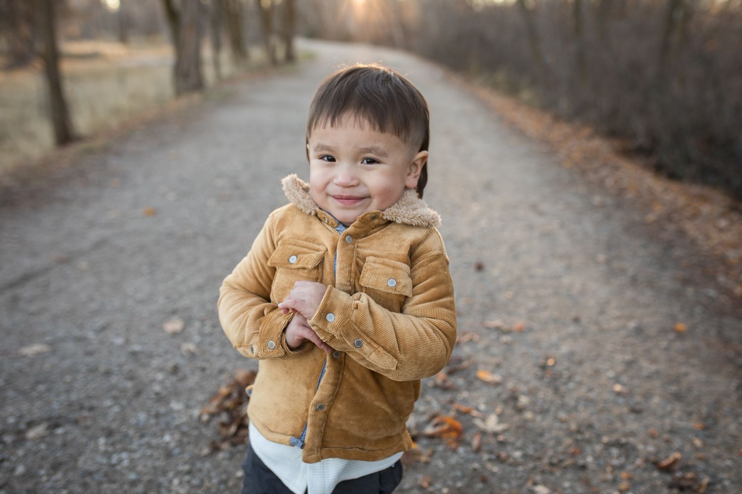 Toddler smiling in a tan jacket on a path at Reid Merrill Park in Eagle, Idaho during golden winter light.