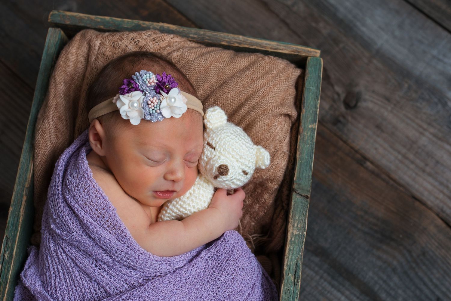 Newborn baby girl wrapped in purple cuddling a small teddy bear during a studio newborn session in Nampa, Idaho.