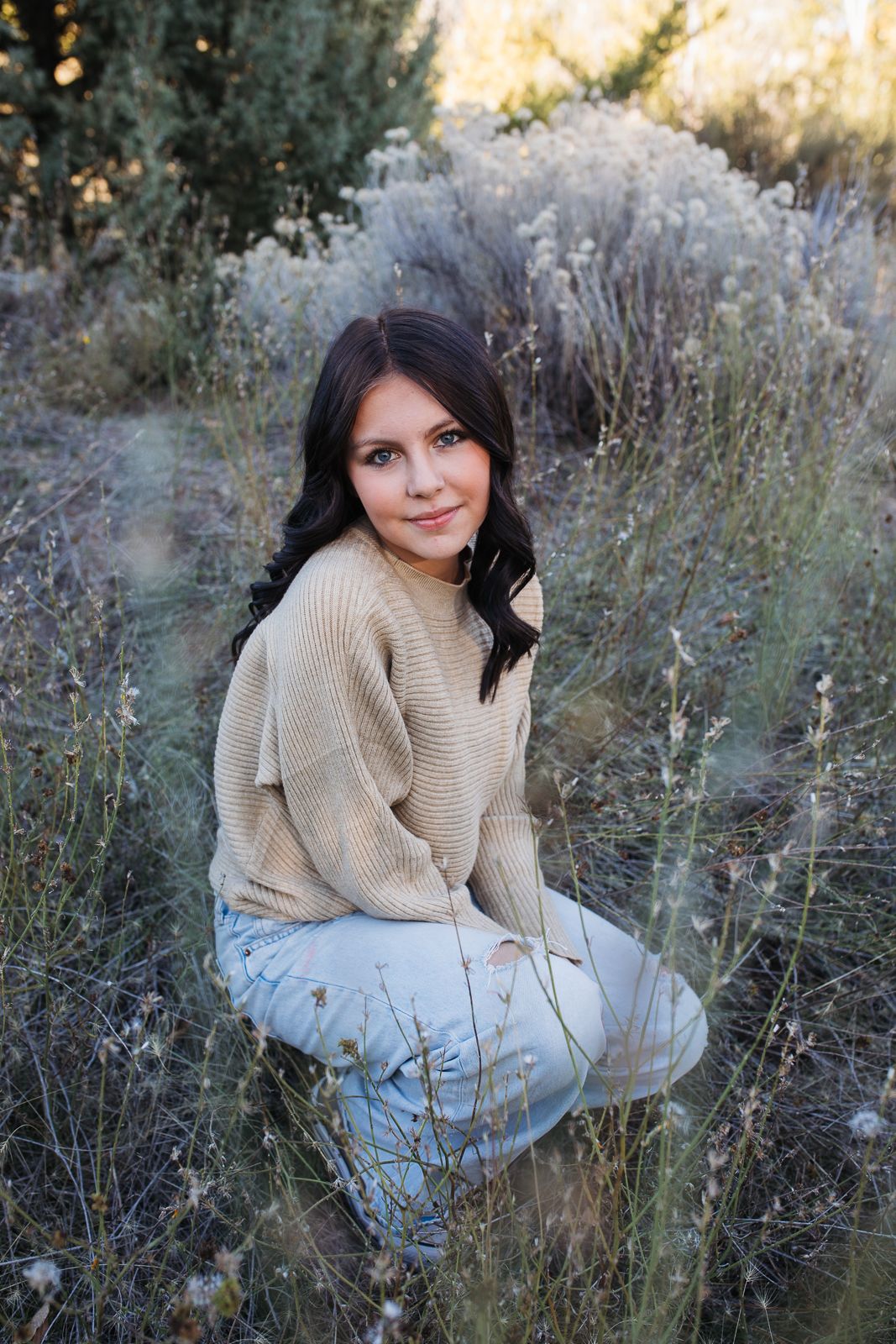 A person wearing a beige sweater sits in a natural outdoor setting among tall grass and wildflowers at sunset. Boise foothills family photog