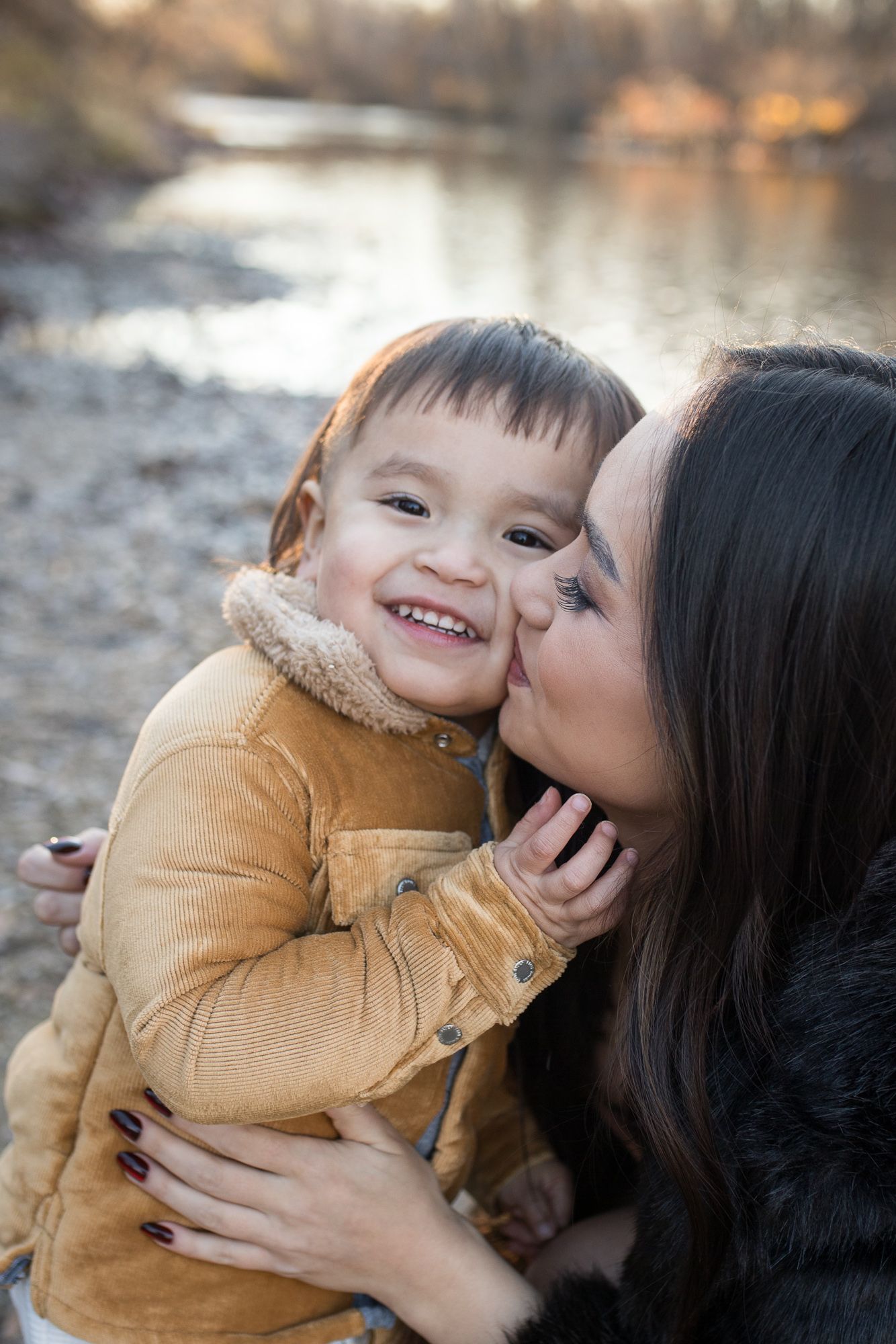 Mother kissing her smiling toddler by the river at Reid Merrill Park in Eagle, Idaho during golden winter light