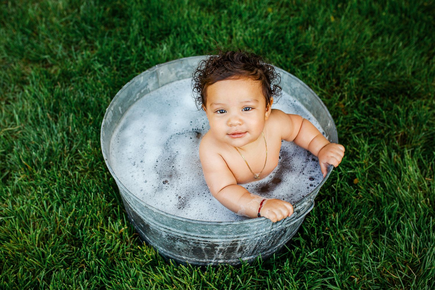 A baby sits in a galvanized metal tub filled with bubbles on green grass during an outdoor bath time - 9 month milestone photos Boise Idaho