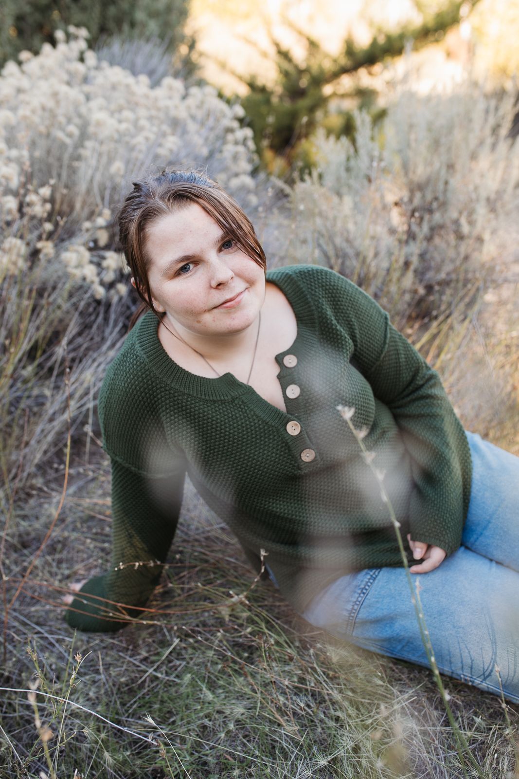 A person in a green sweater and jeans reclines in tall grass during a golden hour outdoor photo session. Boise foothills family photography