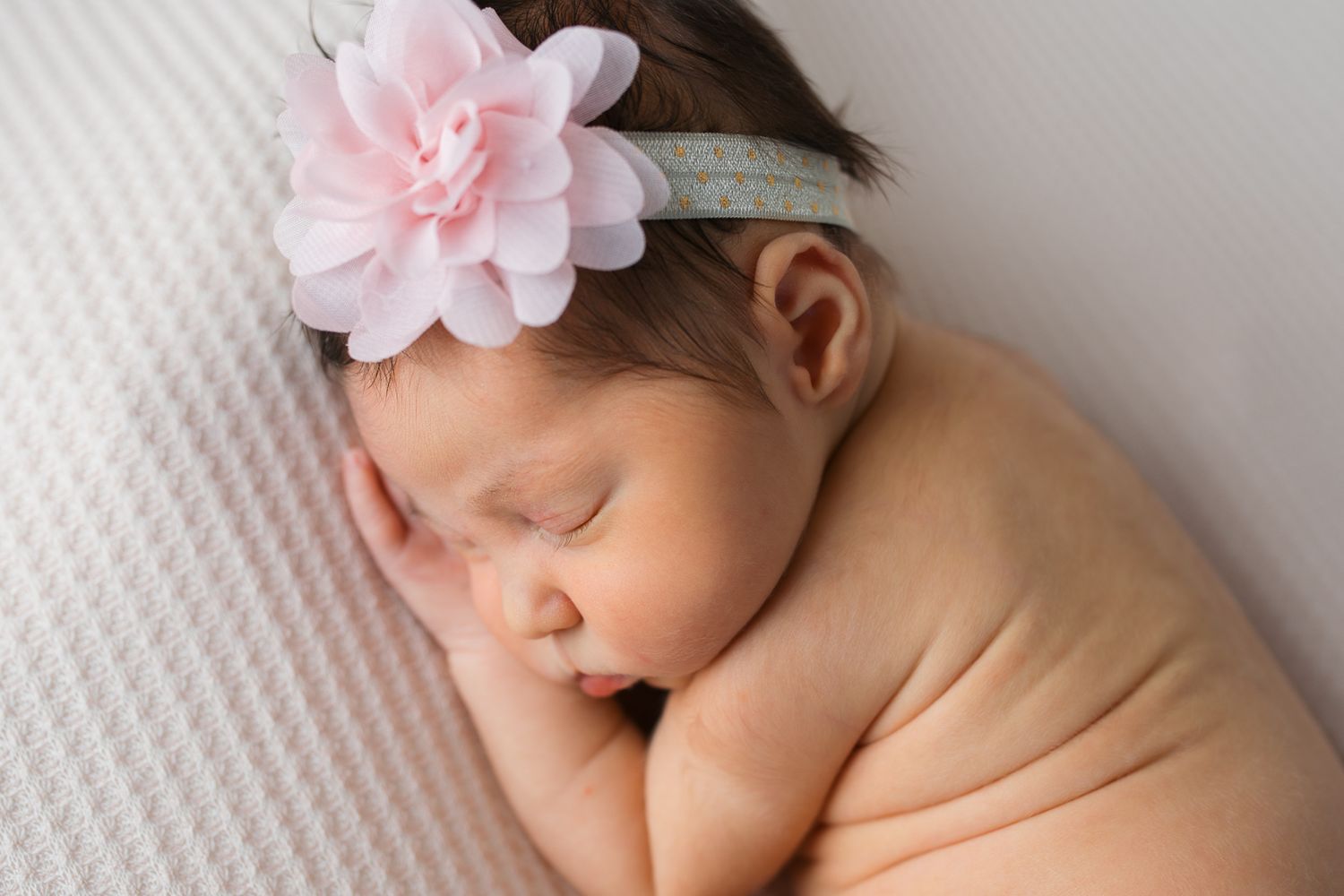 A newborn baby sleeps peacefully wearing a delicate pink flower headband against a white textured background.  studio newborn photography