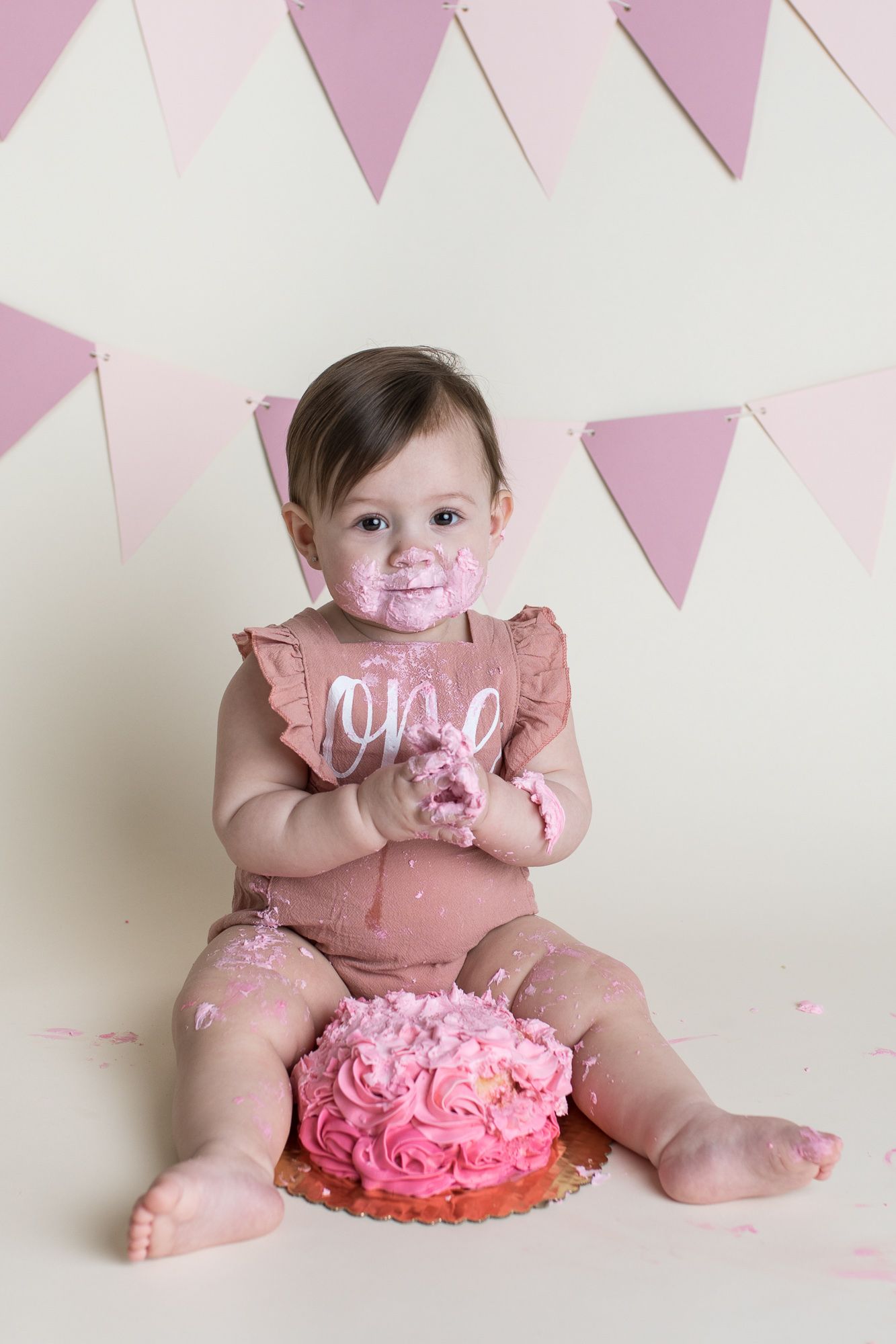 Pink frosted cake smash photo session with pink triangle bunting banner decorations in the background - first birthday photos Boise Idaho
