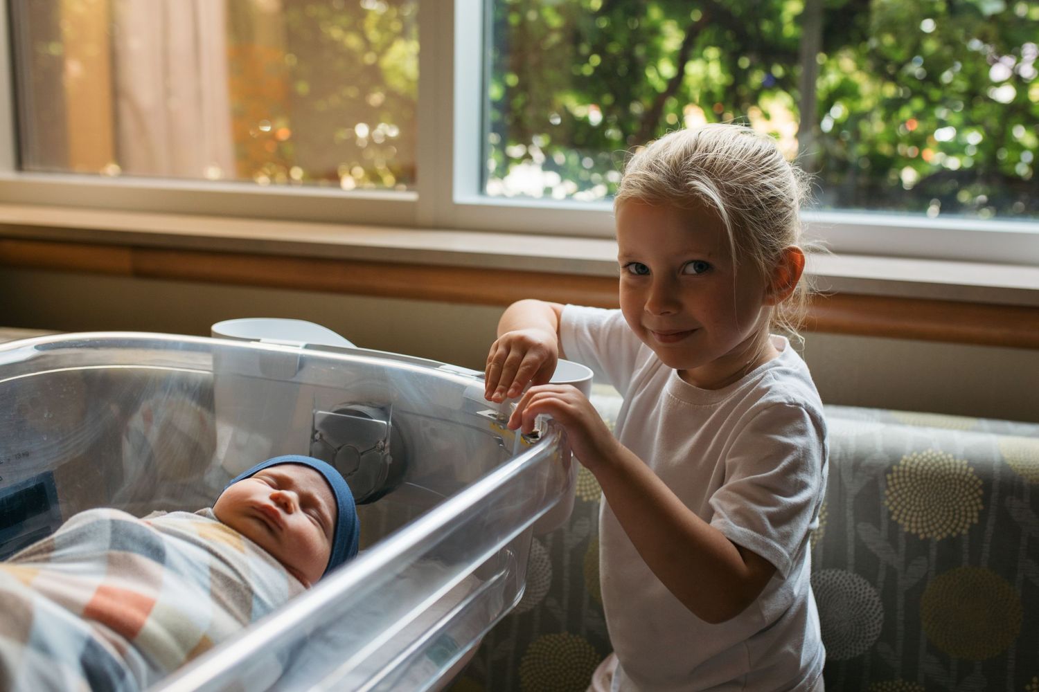 Newborn baby sleeping in a hospital bassinet next to a window, with her older toddler sibling holding the side - Fresh 48 session in Boise