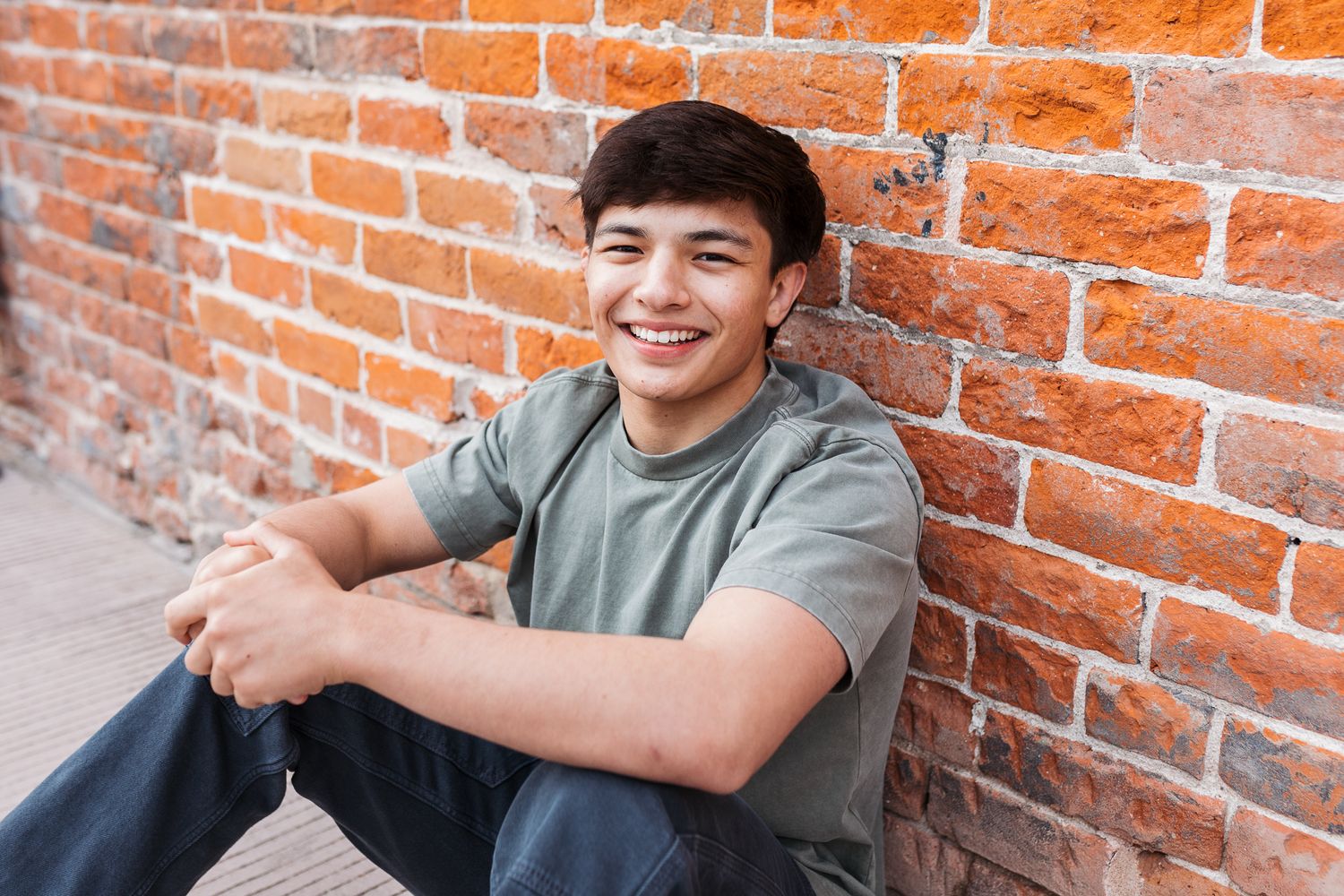 A smiling person in a gray t-shirt sits casually against a rustic red brick wall. - Senior photos Boise Idaho