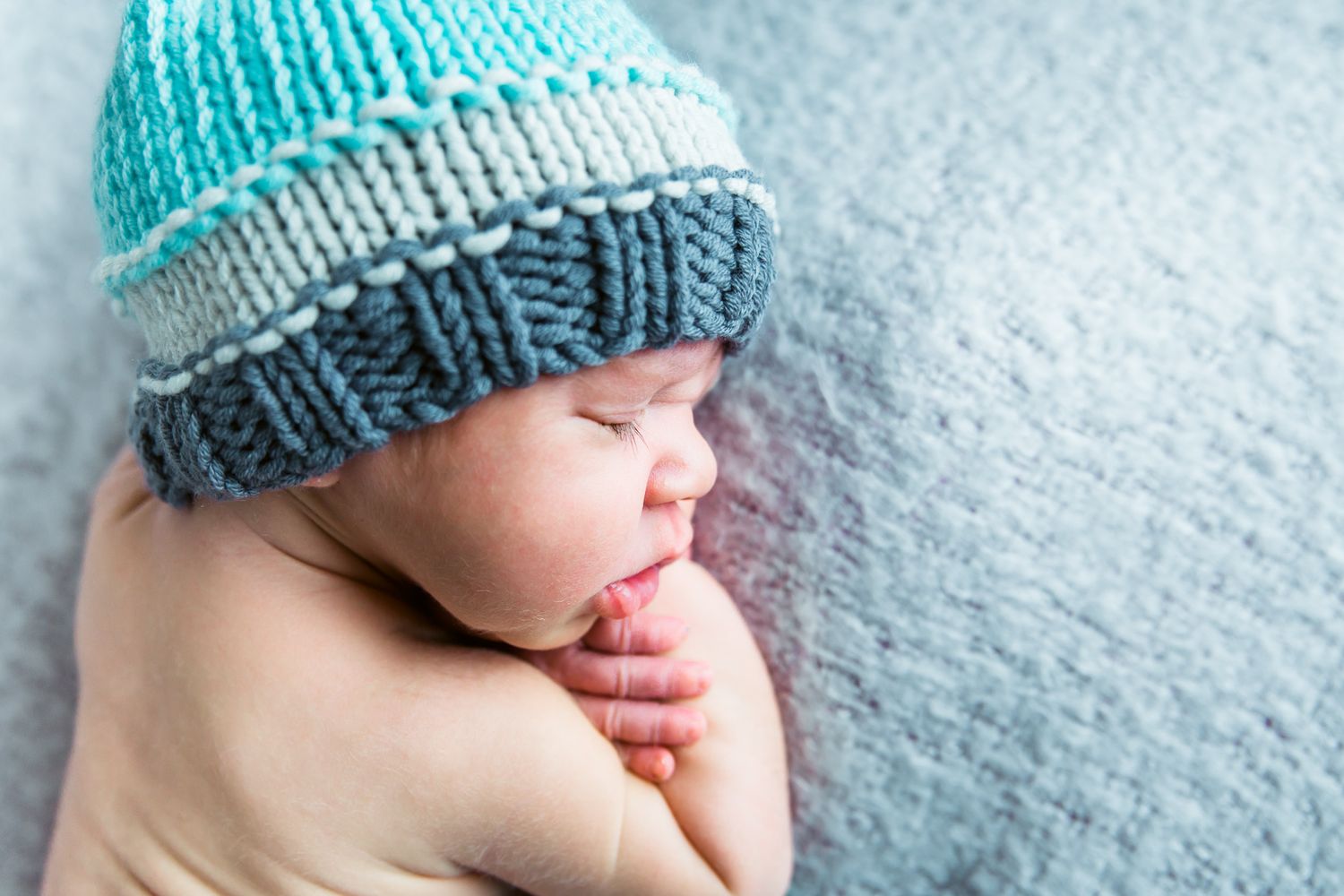 A small baby wearing a knit turquoise and gray beanie sleeps peacefully on a white textured surface studio newborn photography Nampa Idaho