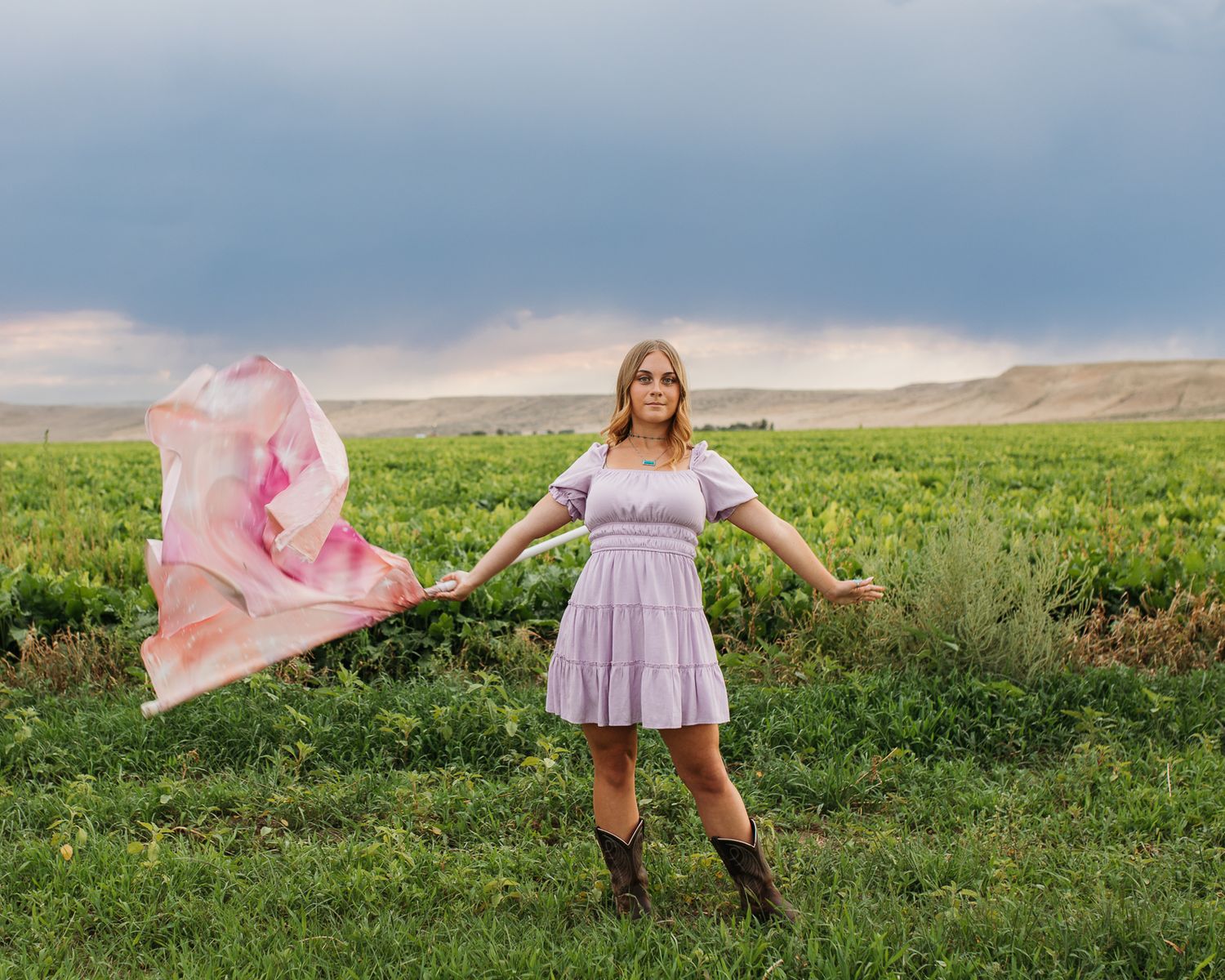 A girl in a flowing pink dress twirls a Color Guard Flag across a lush green meadow with mountains in the background - Senior photos Boise