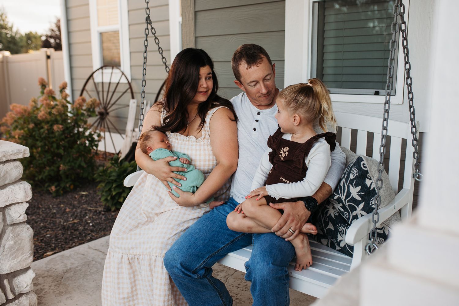 Family sits together on a white porch swing outside their home on a summer evening - lifestyle in-home newborn photos Boise