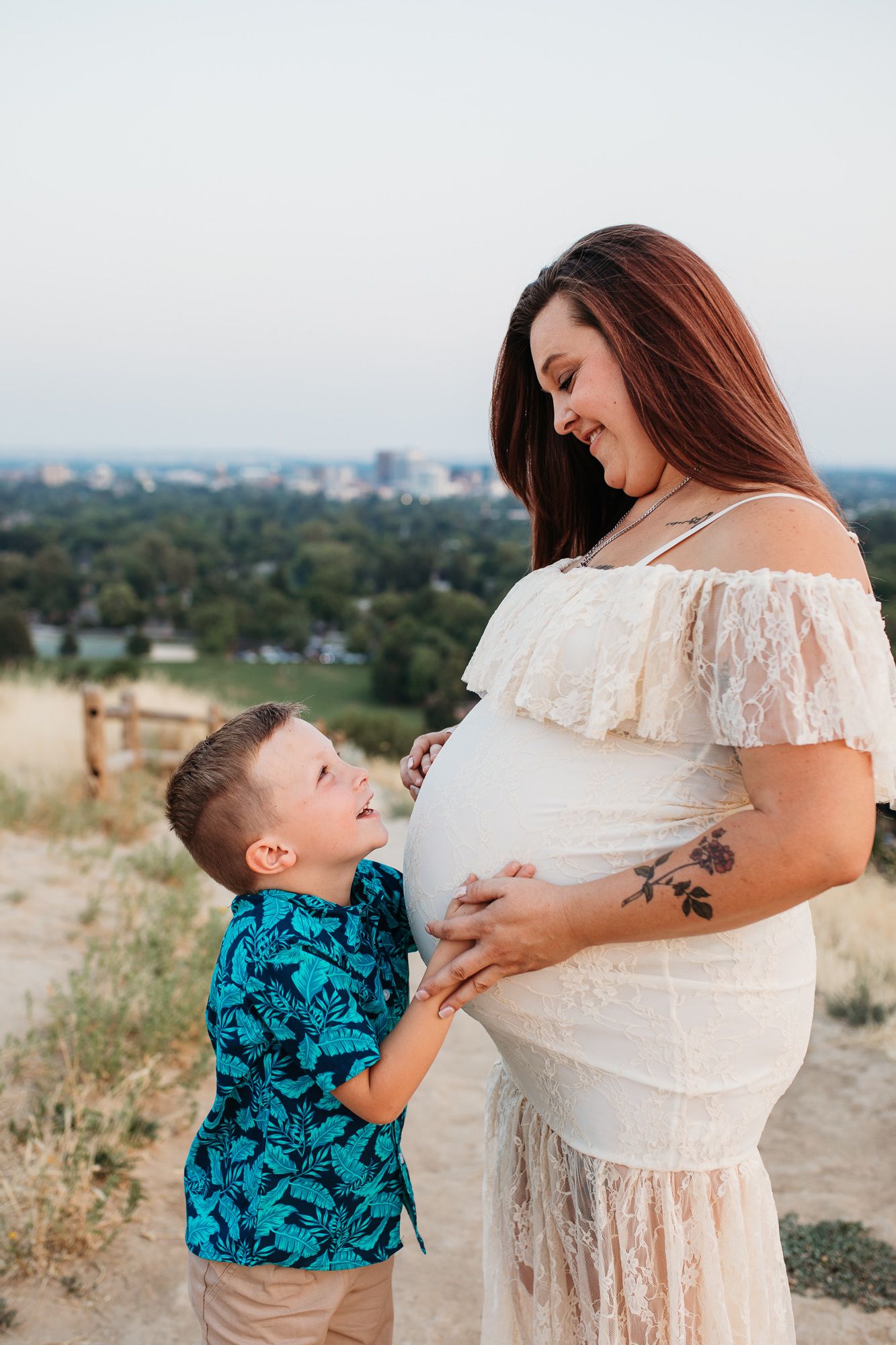 A touching maternity photo series showing a young boy in a blue patterned shirt kissing his mother's pregnant belly outdoors - Boise Idaho