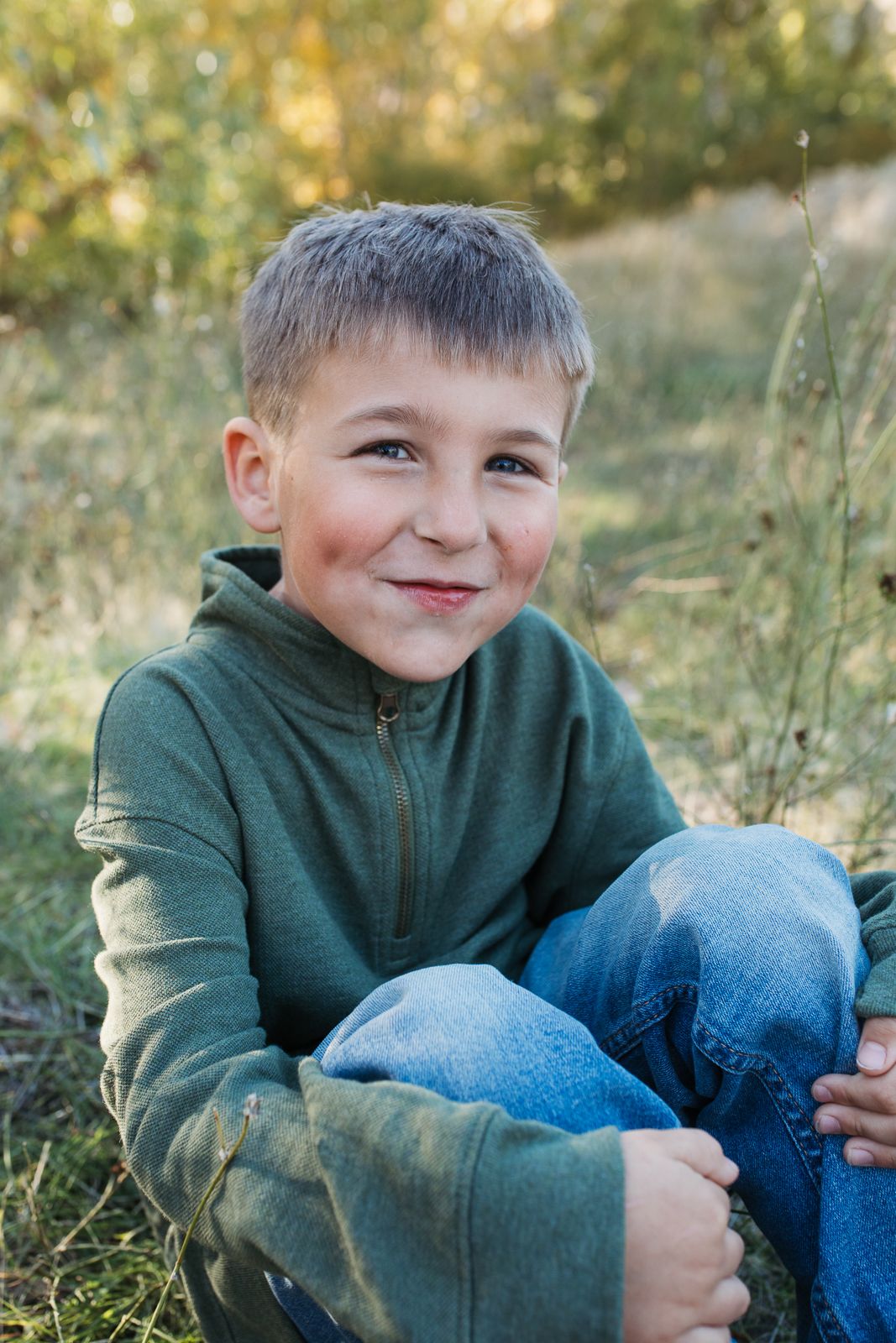 A child in a green sweater sits in tall grass during golden hour lighting with a warm, candid expression. Boise foothills family photography