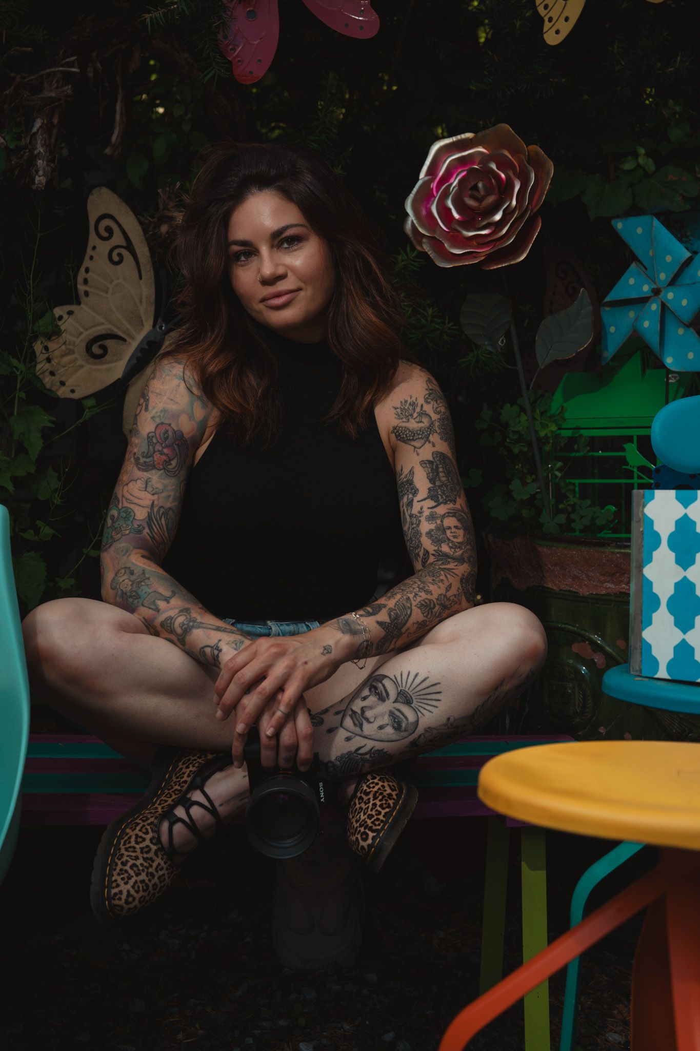 Tattooed woman sitting cross-legged in colorful garden setting with metal flower art and butterfly decorations.