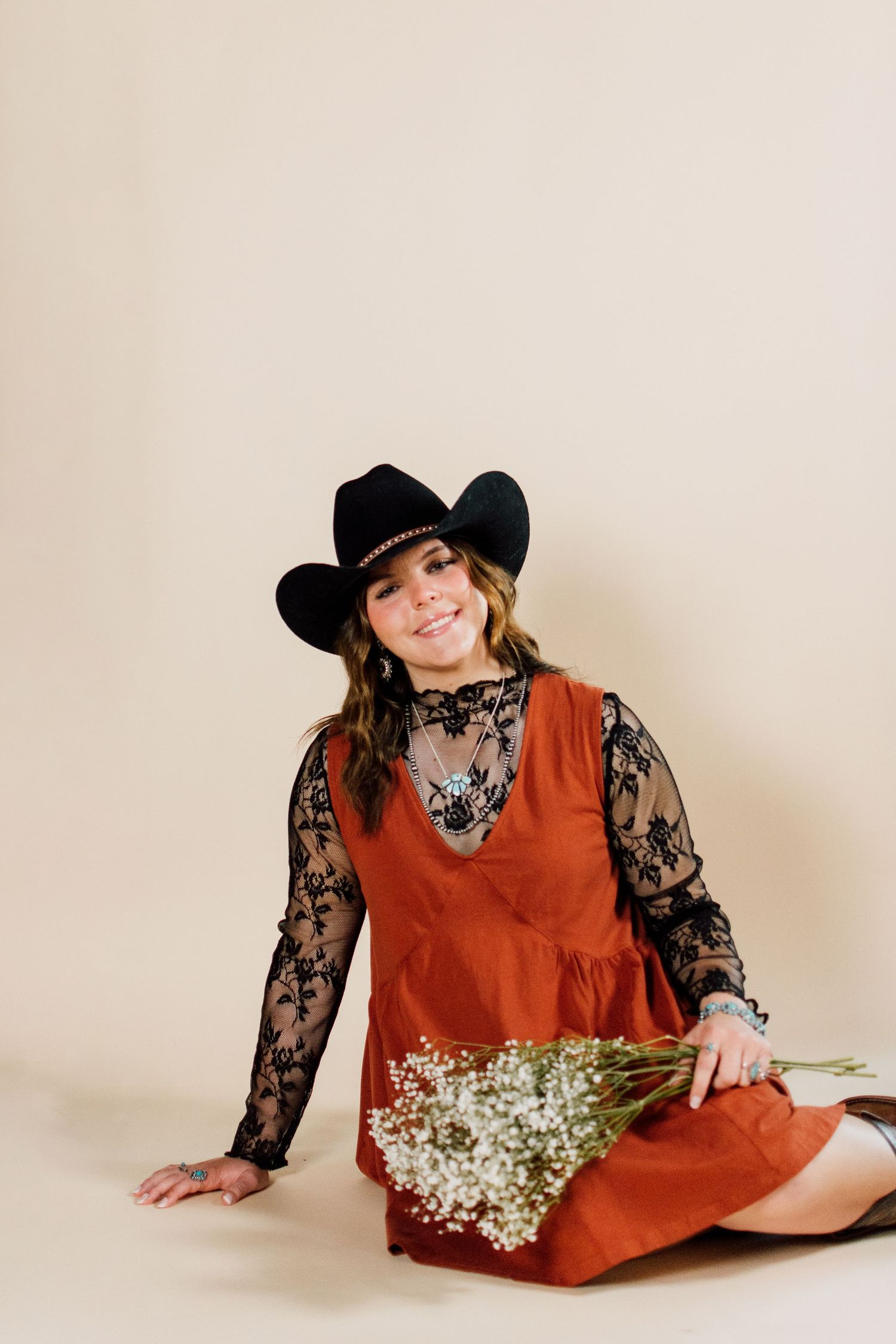 Person in rustic outfit with cowboy hat holds dried flowers against beige background.