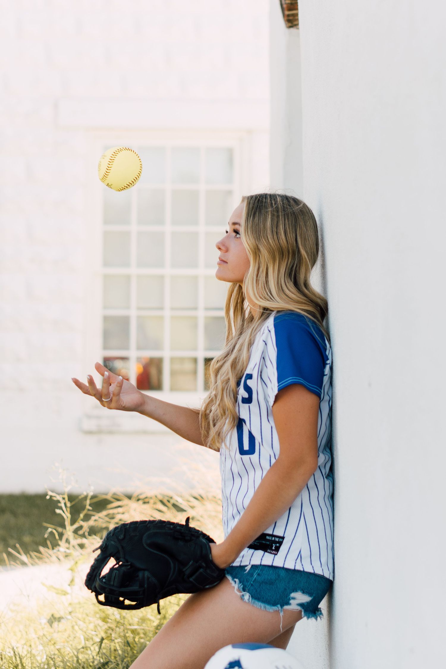 Softball player in blue and white striped uniform tosses ball while sitting against white wall.