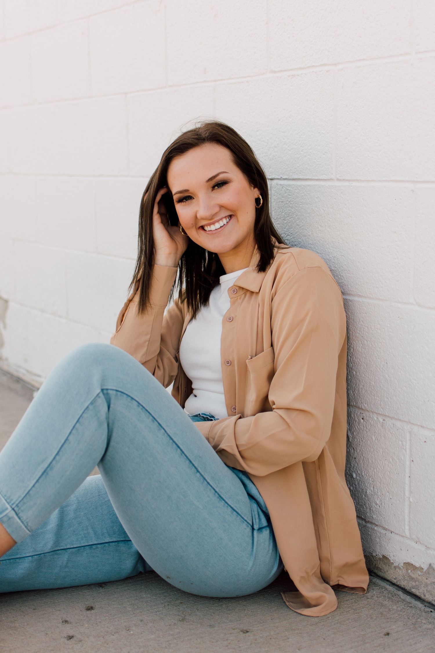 Casual portrait in light jacket and jeans sitting against white wall.