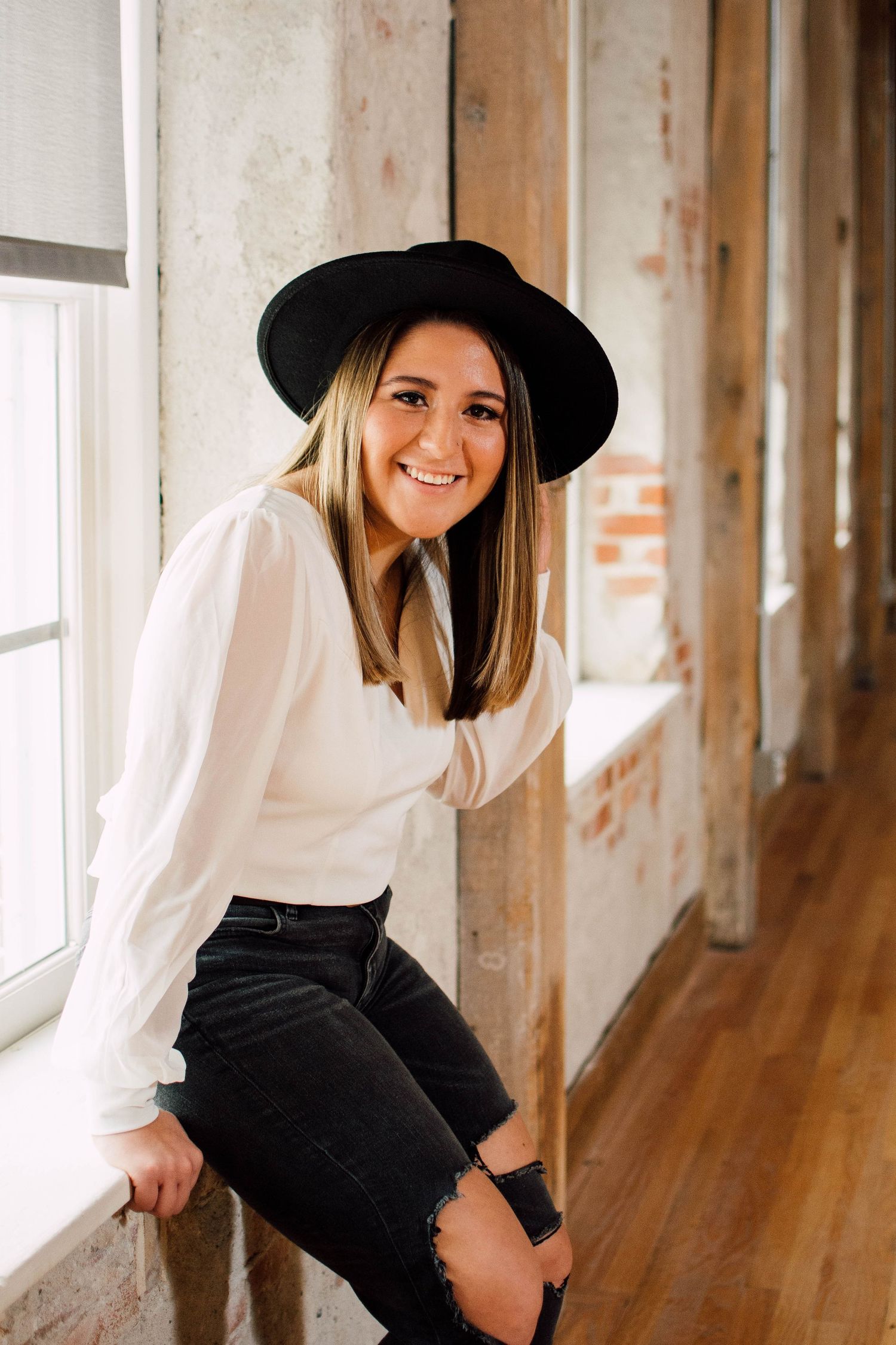 Woman in white blouse and black pants poses by window in rustic interior setting.