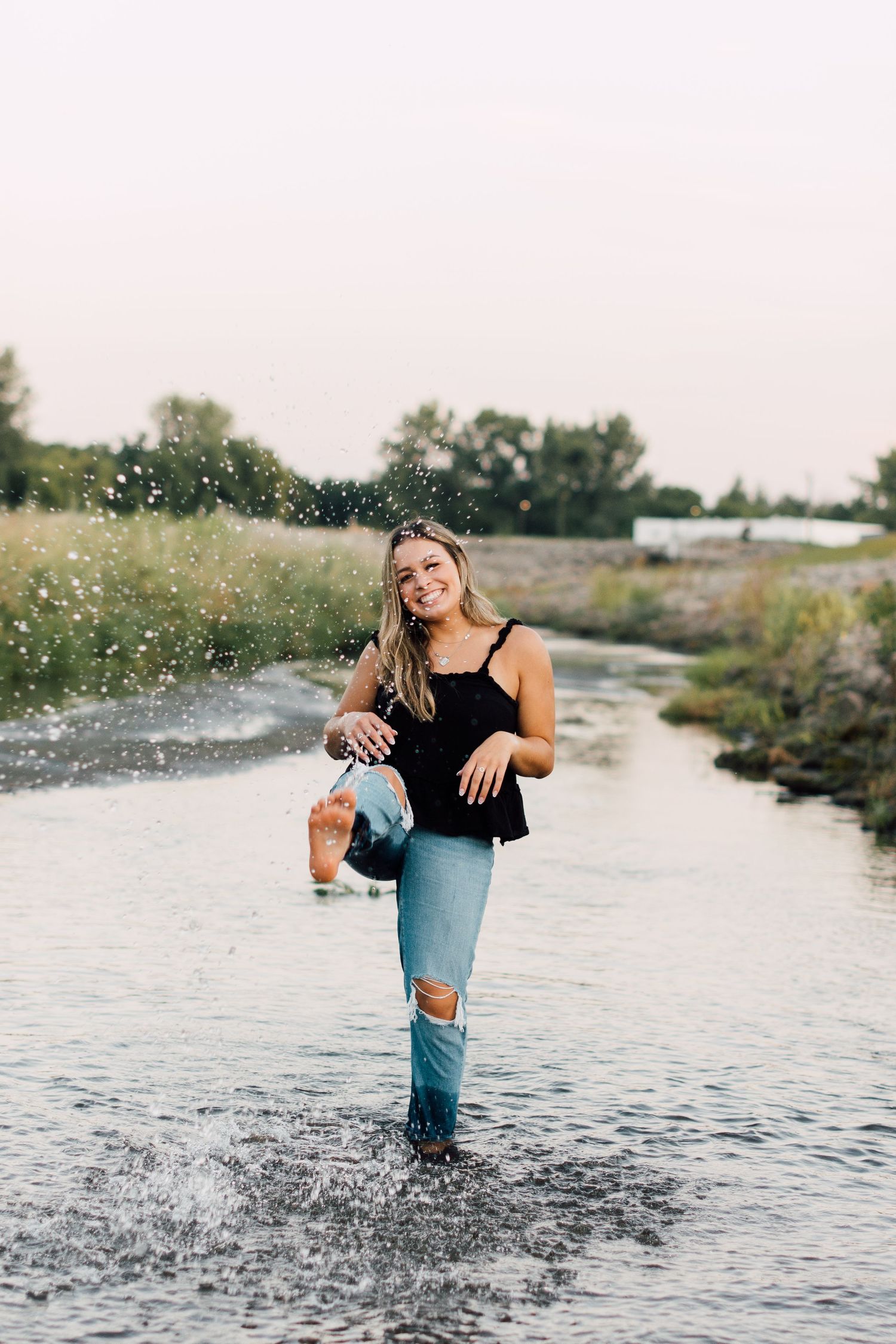Person splashing in shallow water wearing ripped jeans and black top during sunset photoshoot.