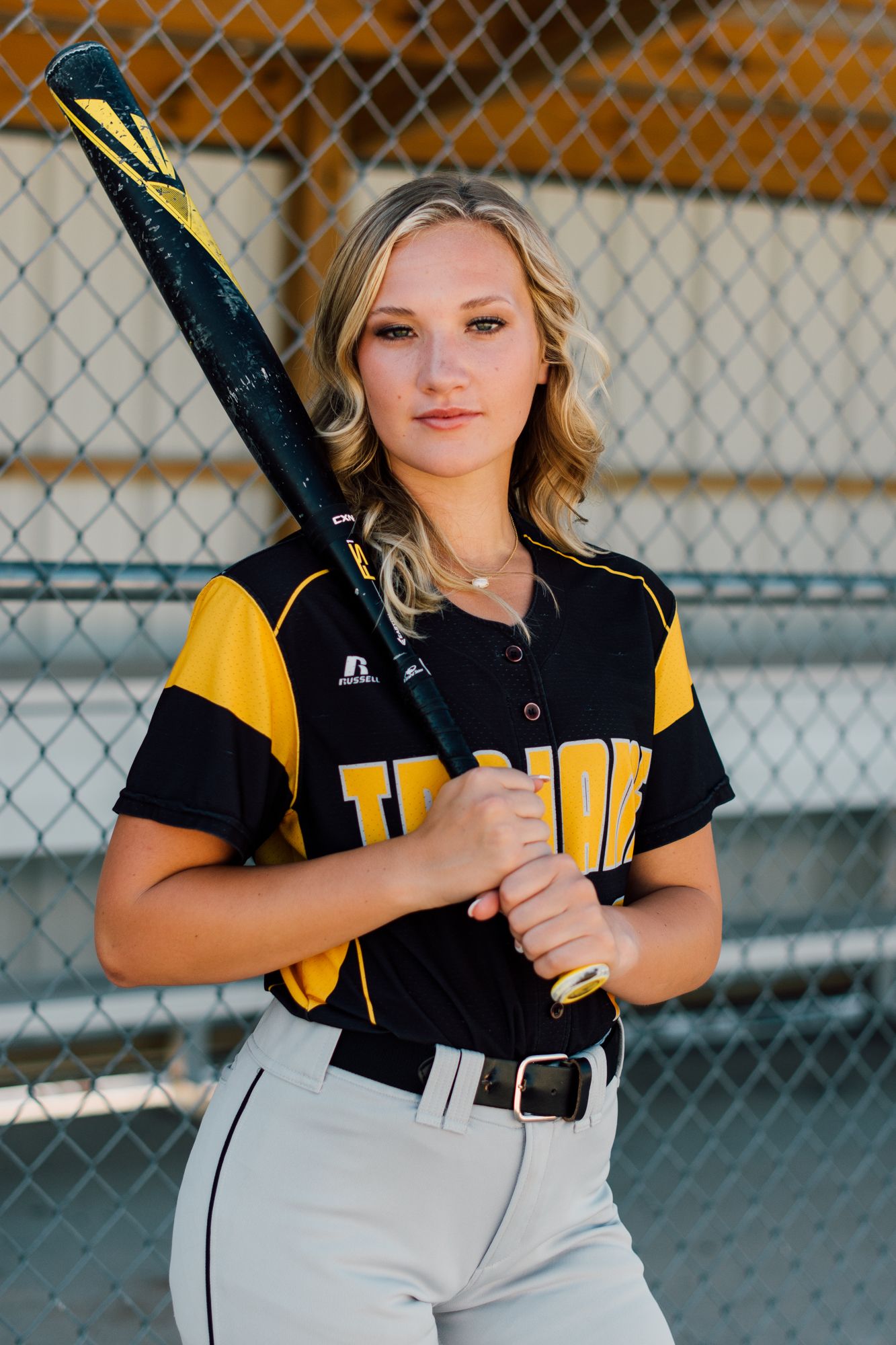 Softball player poses in black and yellow uniform with bat against chain link fence.