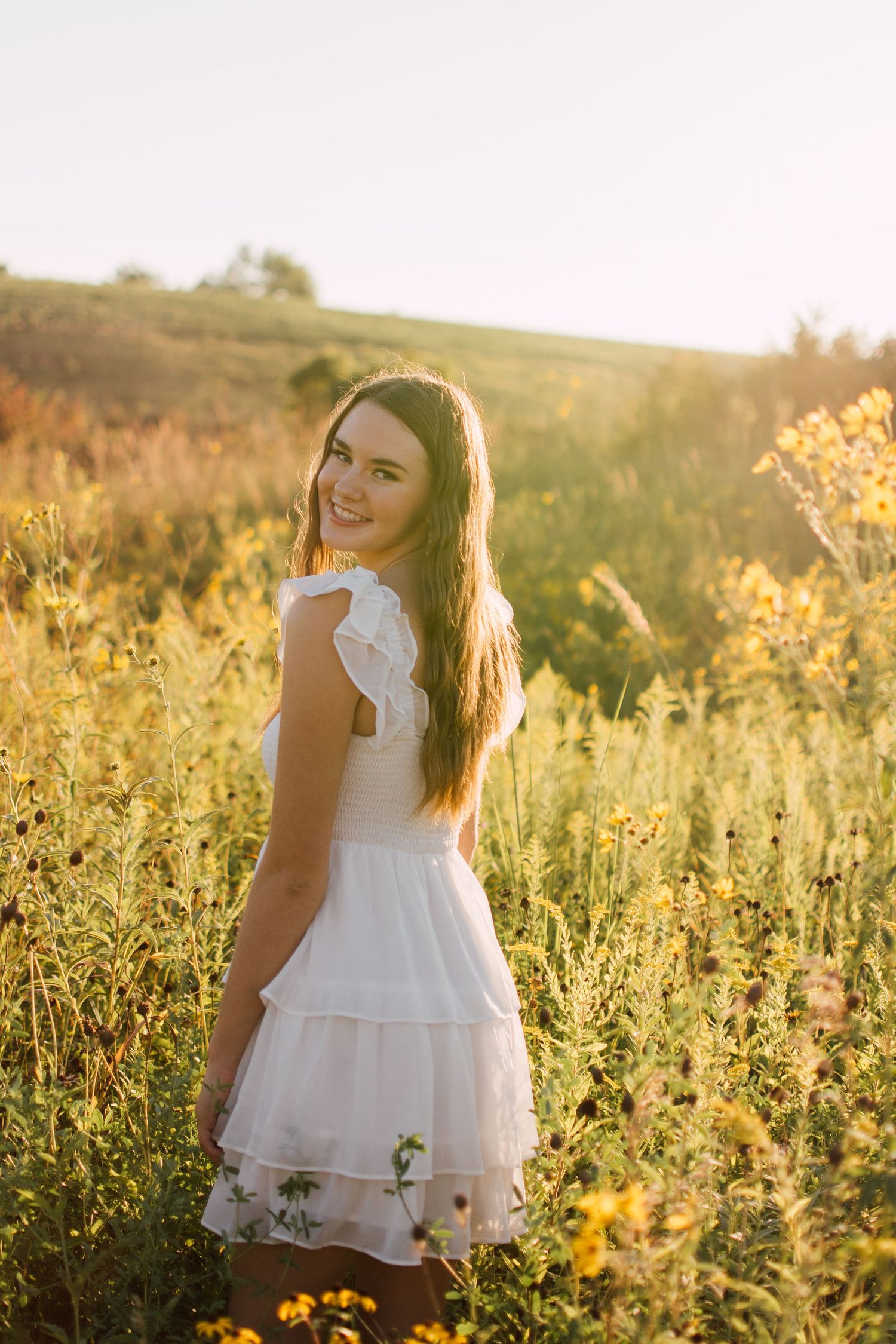 Woman in white dress poses in golden sunlit field of wildflowers at sunset.