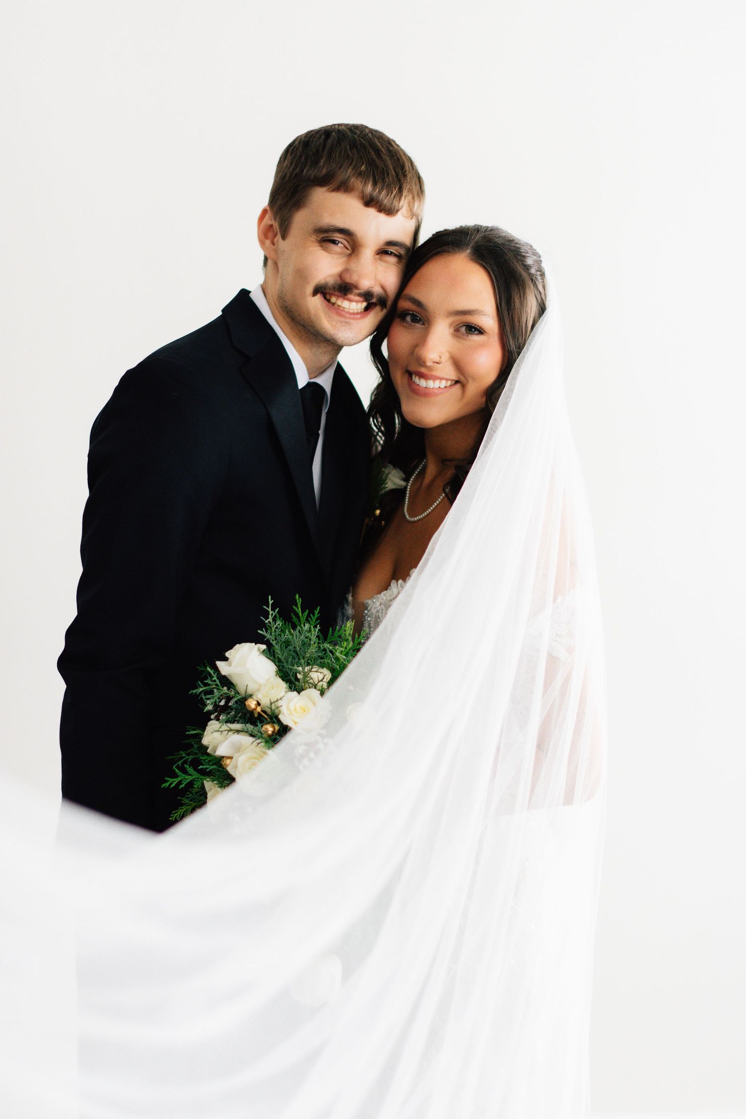Intimate wedding portrait with flowing white veil against bright studio background.