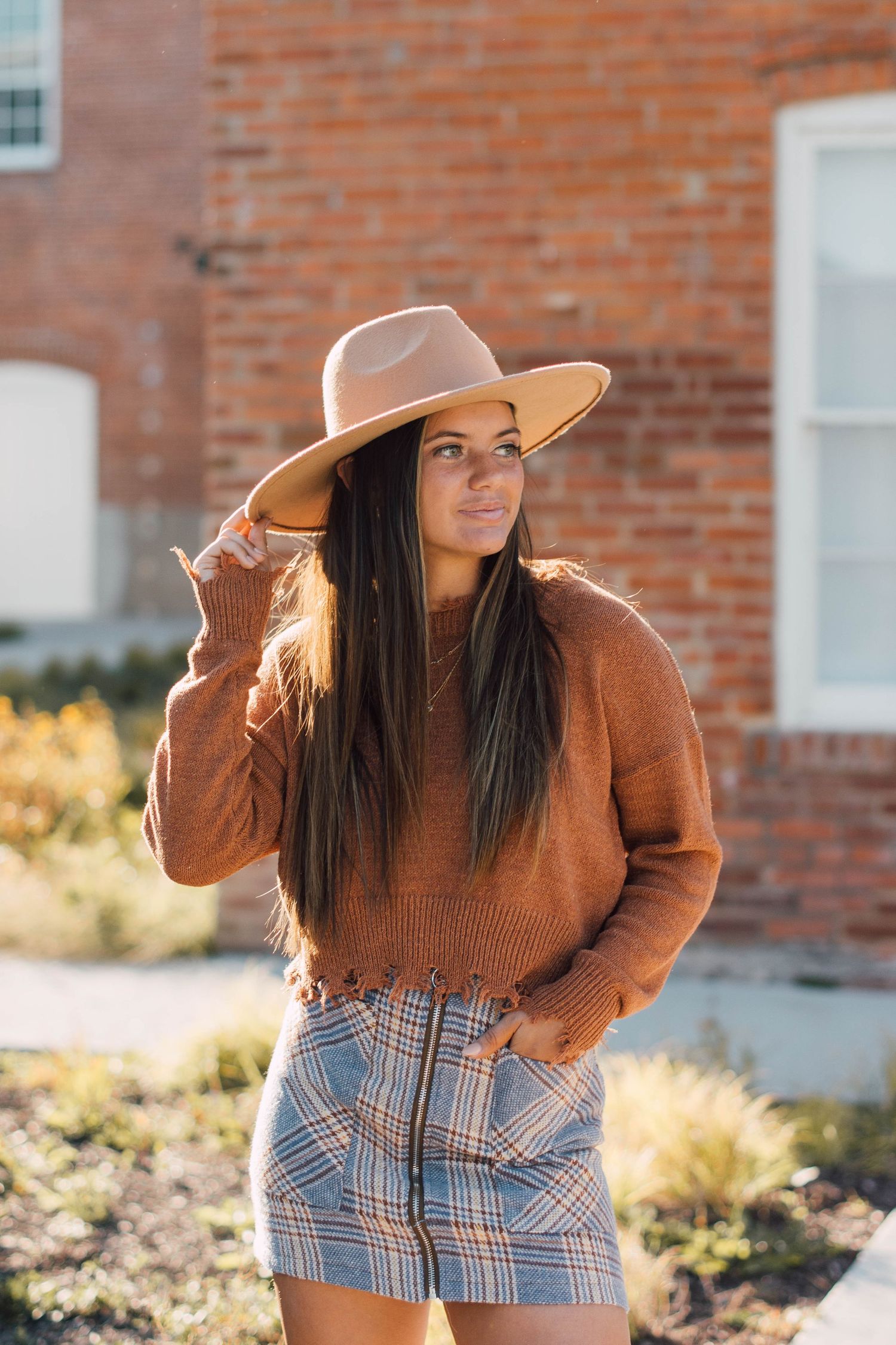 Fashion model in brown sweater and plaid pants poses against brick wall in autumn sunlight.