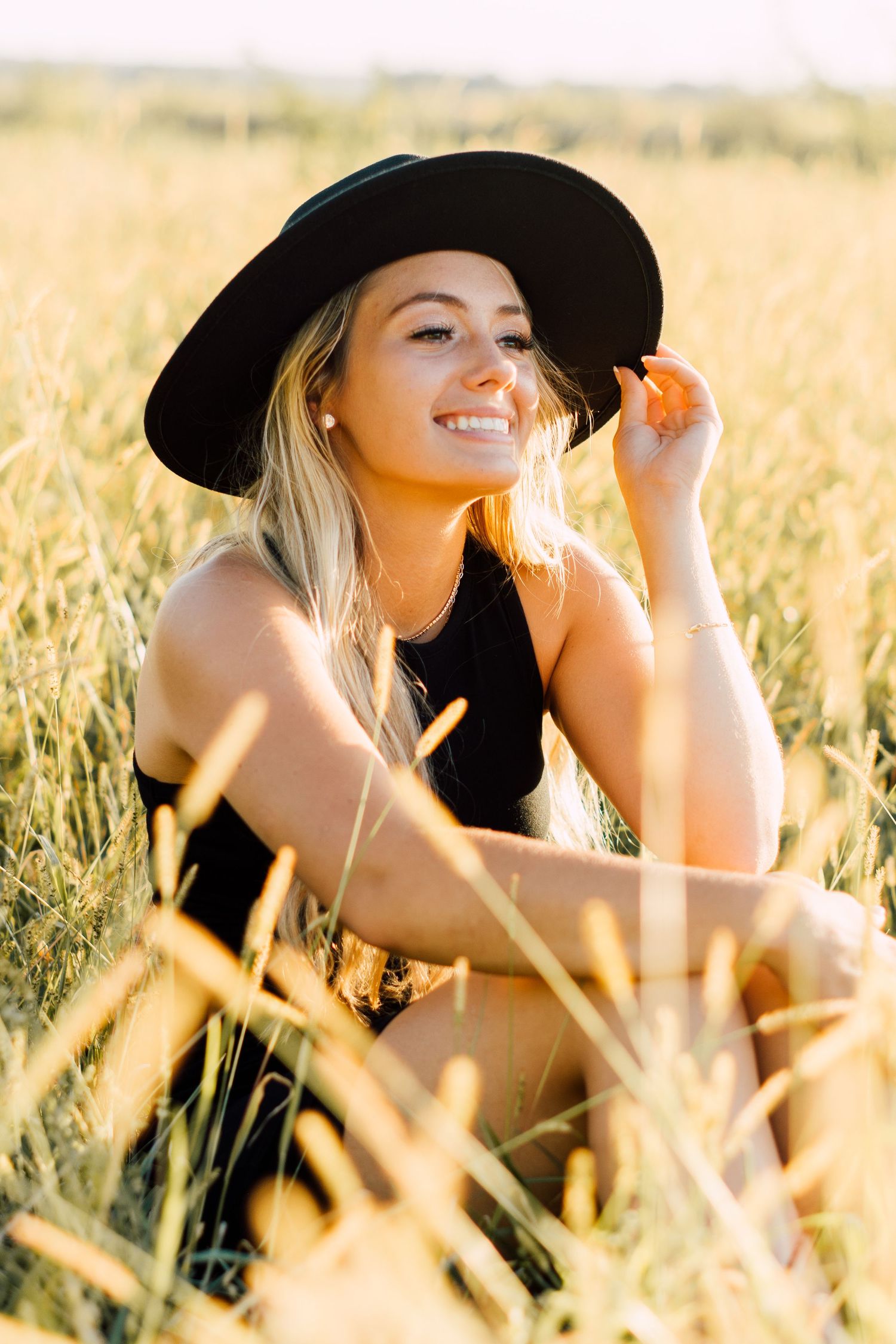 Artistic portraits in golden wheat field wearing wide-brim black hat at sunset.
