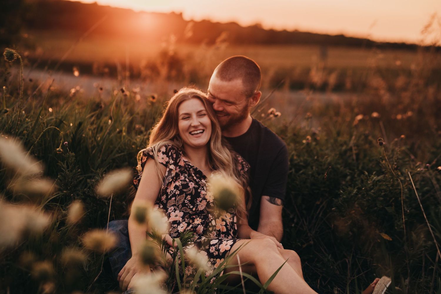 A couple shares an intimate moment in a wildflower field at sunset while sitting among tall grass and flowers.