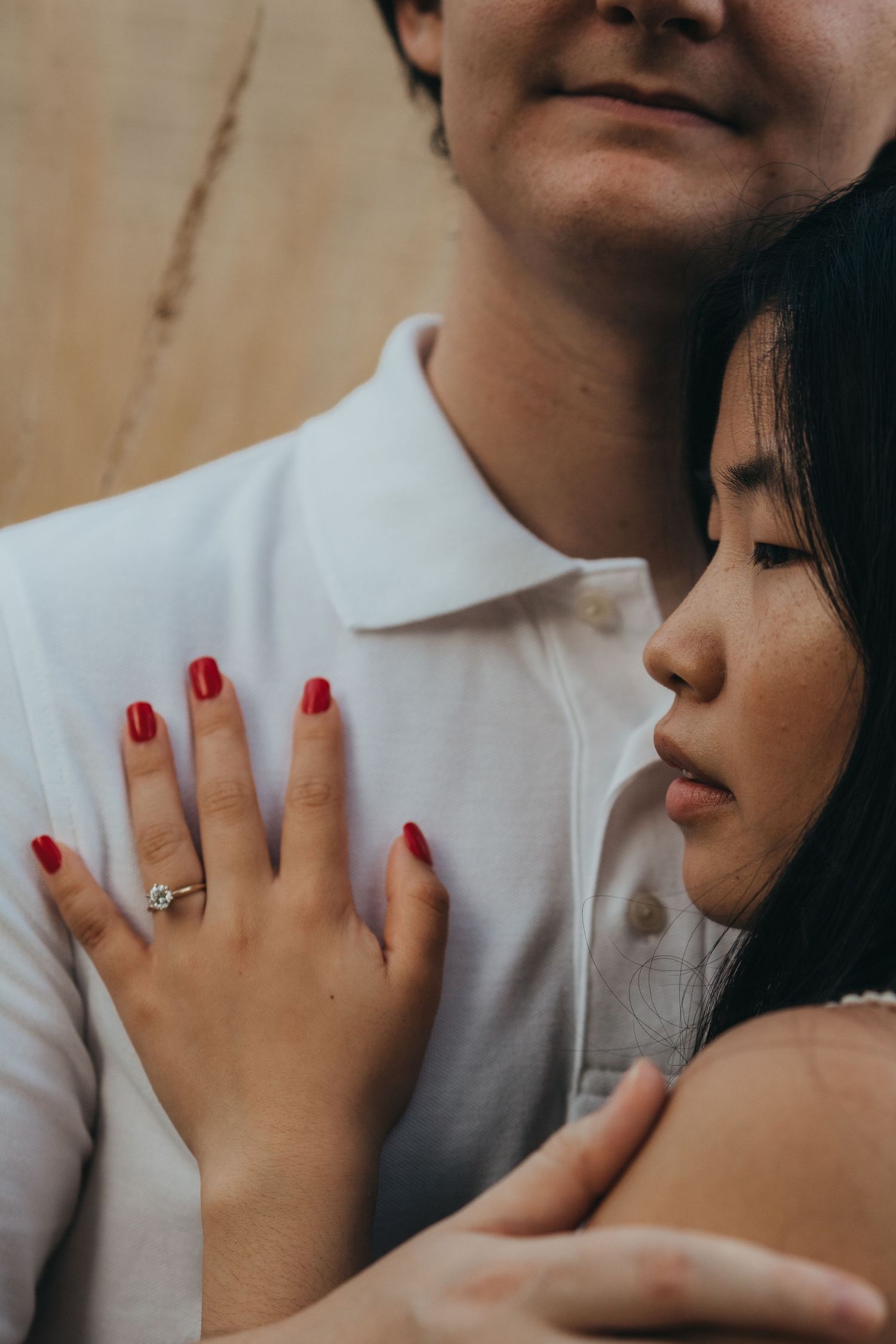 Close-up intimate portrait showing a couple embracing, with red painted fingernails and an engagement ring visible.