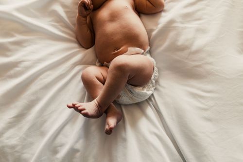 Baby wearing a cloth diaper lies on white bedding with soft brown plush toy nearby.  professional Family photos vigo county