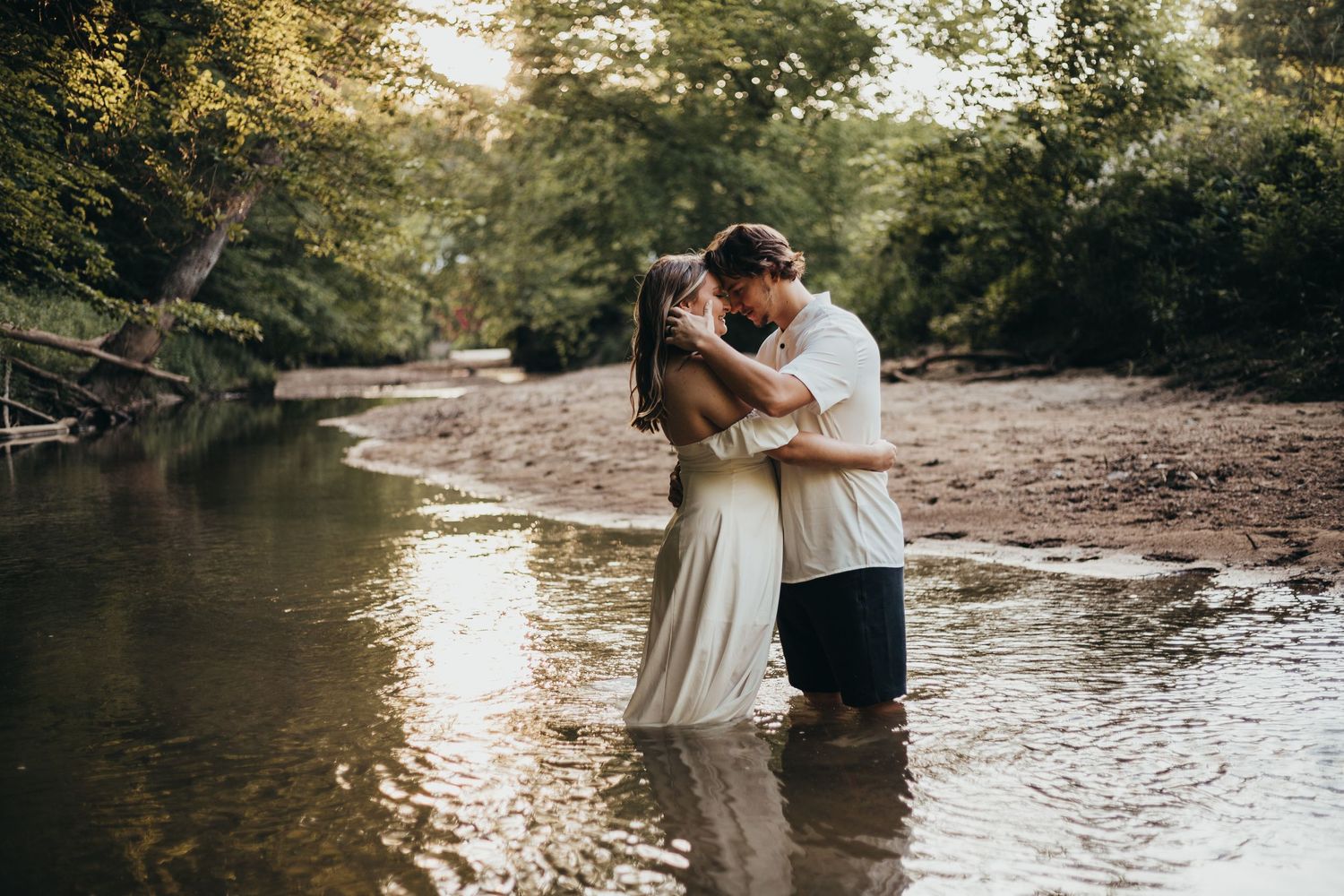 A romantic couple embraces while standing in a shallow creek surrounded by lush forest scenery. couple photography session near parke county