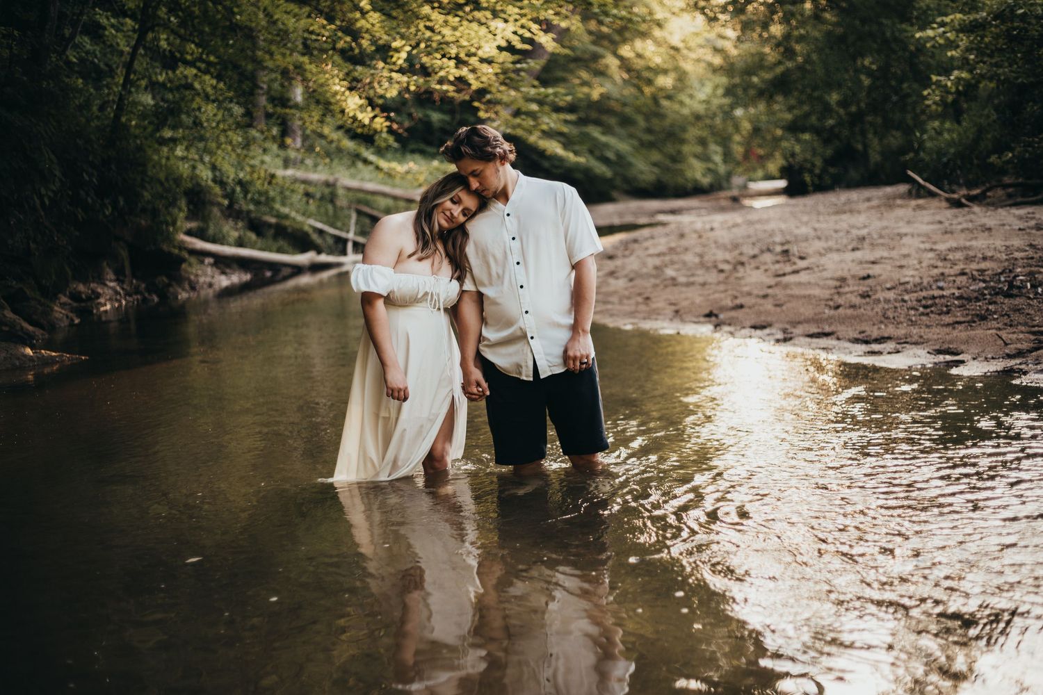 A romantic couple stands in shallow river water surrounded by lush forest during golden hour.