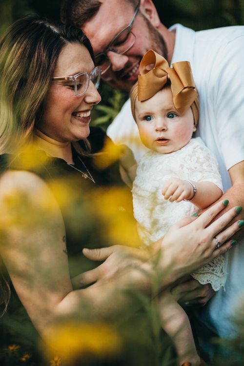 An intimate family moment captured outdoors with parents and baby wearing a gold bow, surrounded by yellow flowers. Family photos vigo count