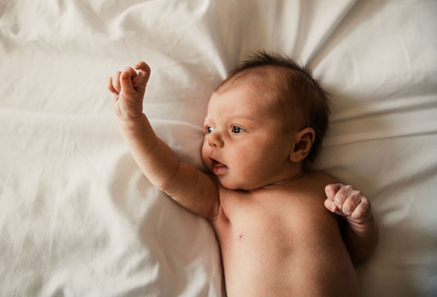 A small baby lying awake on white bedding with one arm raised while resting.