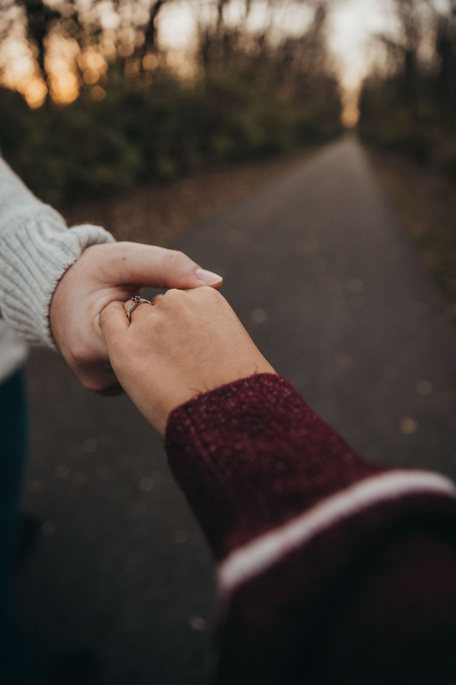 Two hands holding each other on a path at sunset, wearing burgundy and gray sweater sleeves.