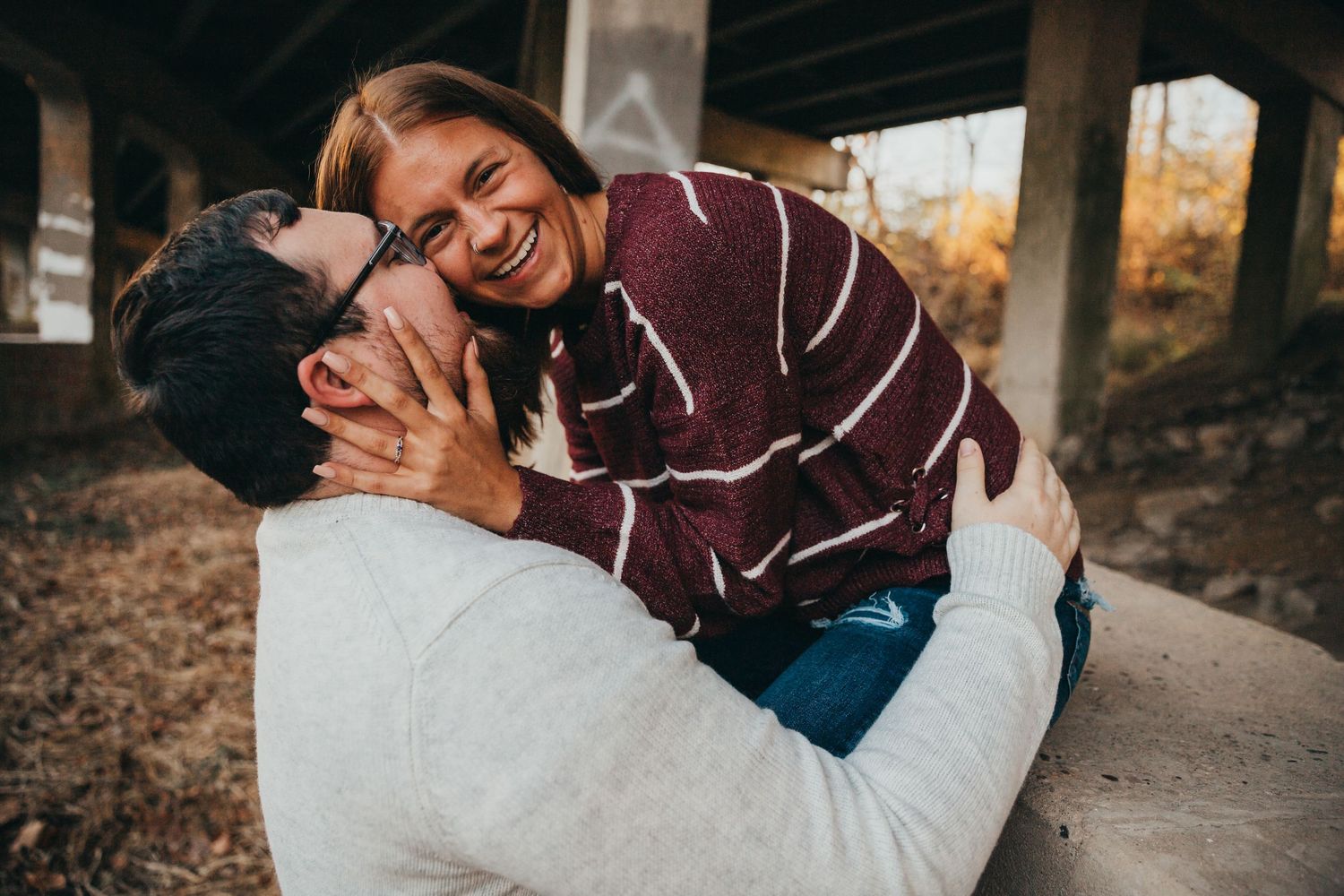 A couple shares a joyful embrace while sitting outdoors during autumn with trees in the background.
