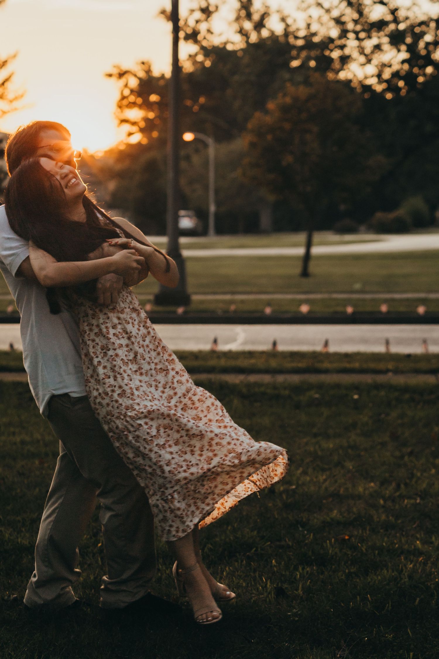 A couple embraces and dances in a park at sunset while wearing summer attire on a warm evening.