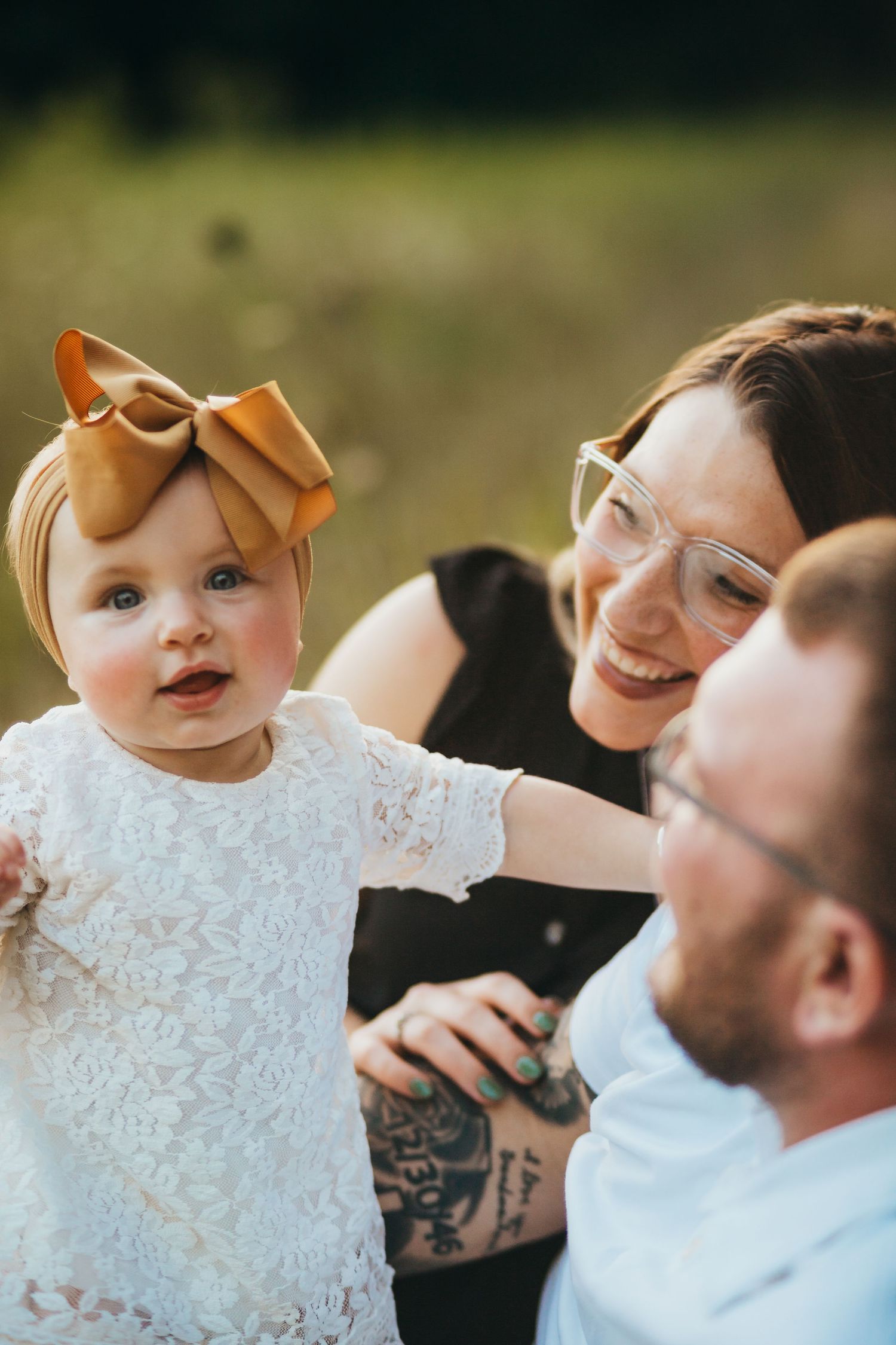 Adorable baby in white lace dress with bow headband sharing sweet moment with parents outdoors.