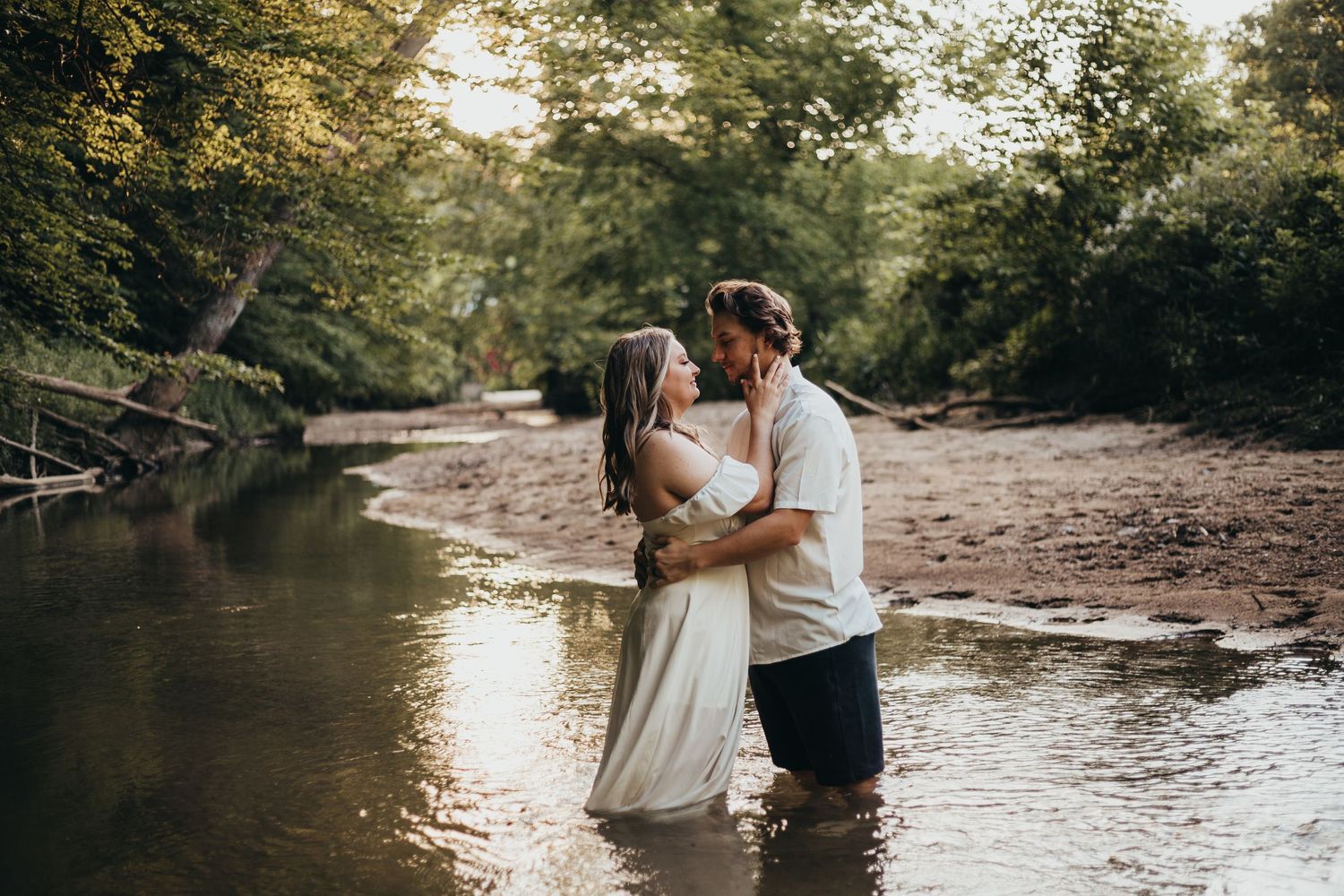 A couple shares an intimate moment while standing in a shallow creek surrounded by lush trees during golden hour.
