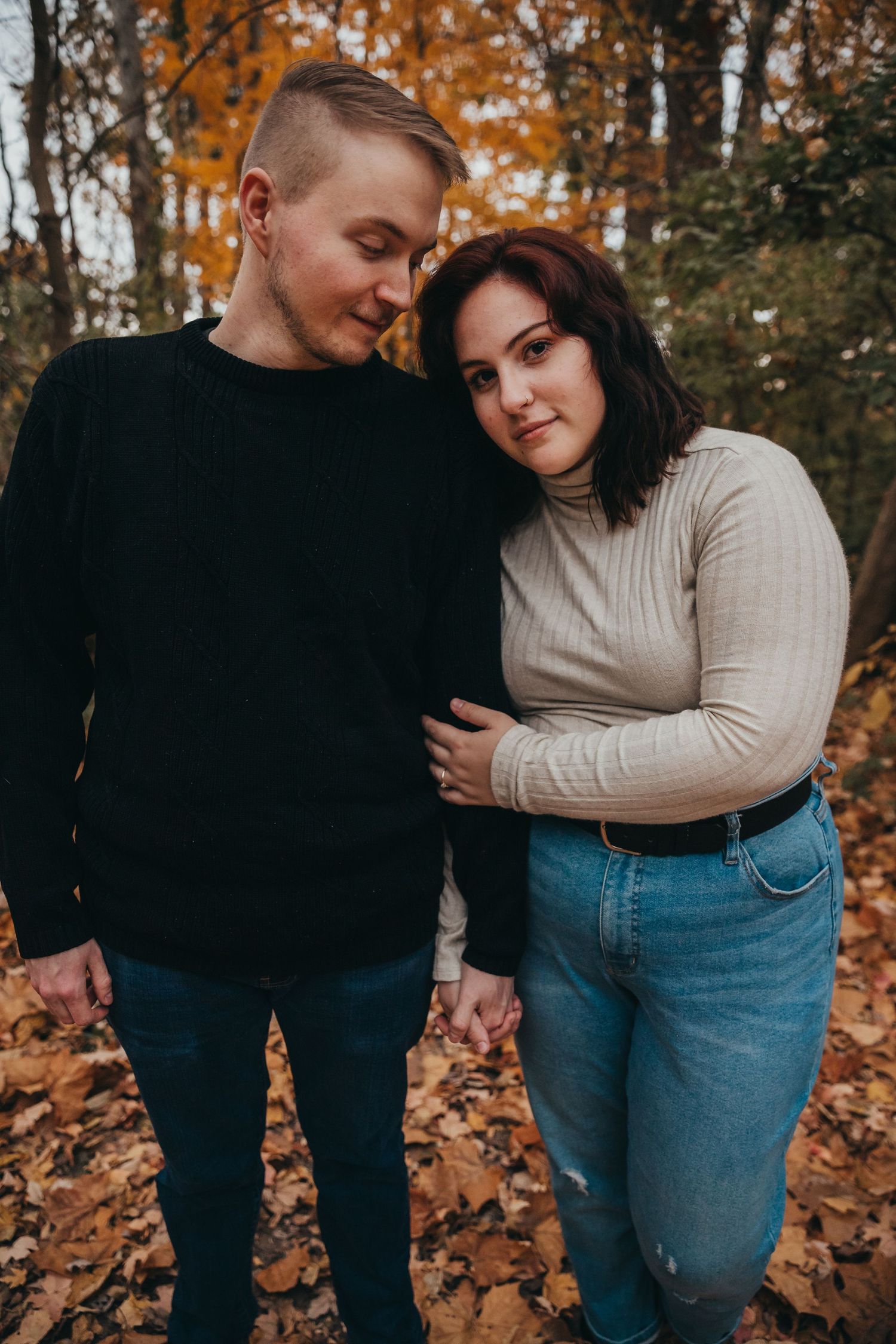 Couple in casual sweaters embracing outdoors among fallen autumn leaves in a romantic forest photoshoot.