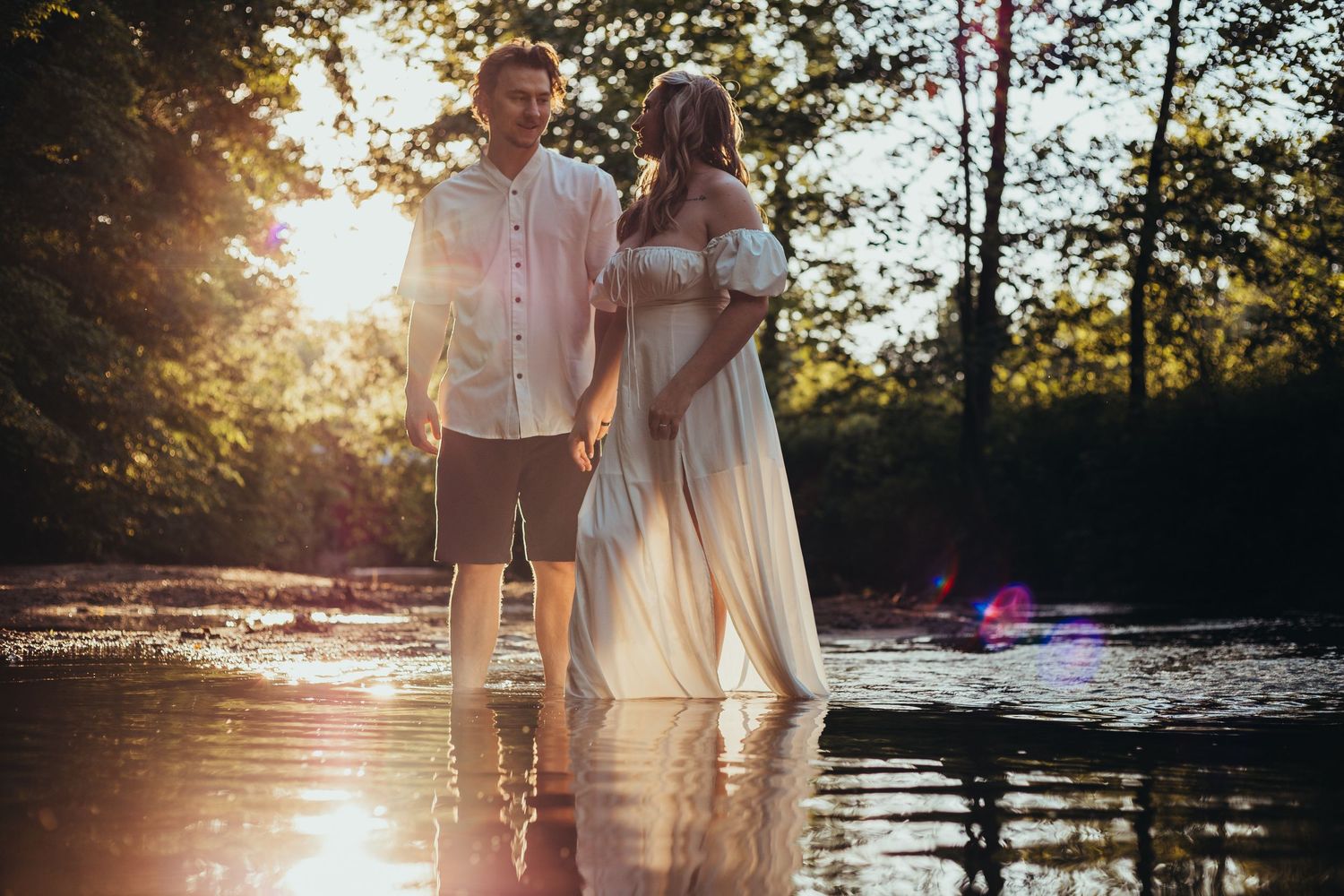 Two people in light clothing embrace while standing in shallow water as sunlight streams through trees.